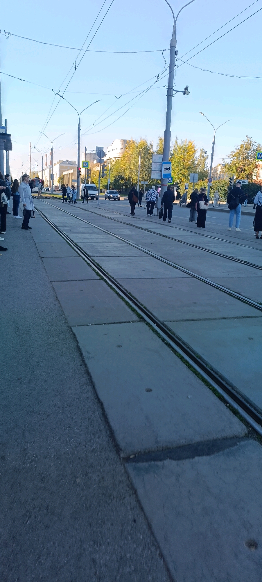 The image depicts a street scene in Perm, Russia, likely in the daytime under a clear sky. The foreground shows a section of paved road adjacent to tram tracks embedded in concrete slabs. The tram tracks run parallel and extend into the distance.
Several people are visible on the sidewalk and crossing the street, some waiting near a bus stop sign. The individuals are dressed in casual attire, and some are carrying bags. There are also vehicles present, including cars and what appears to be a white van. In the background, buildings and trees with autumn foliage can be seen, suggesting the season is likely fall. Streetlights and overhead wires for the tram system are prominent features of the urban infrastructure. The overall impression is of a typical urban environment with everyday activity.