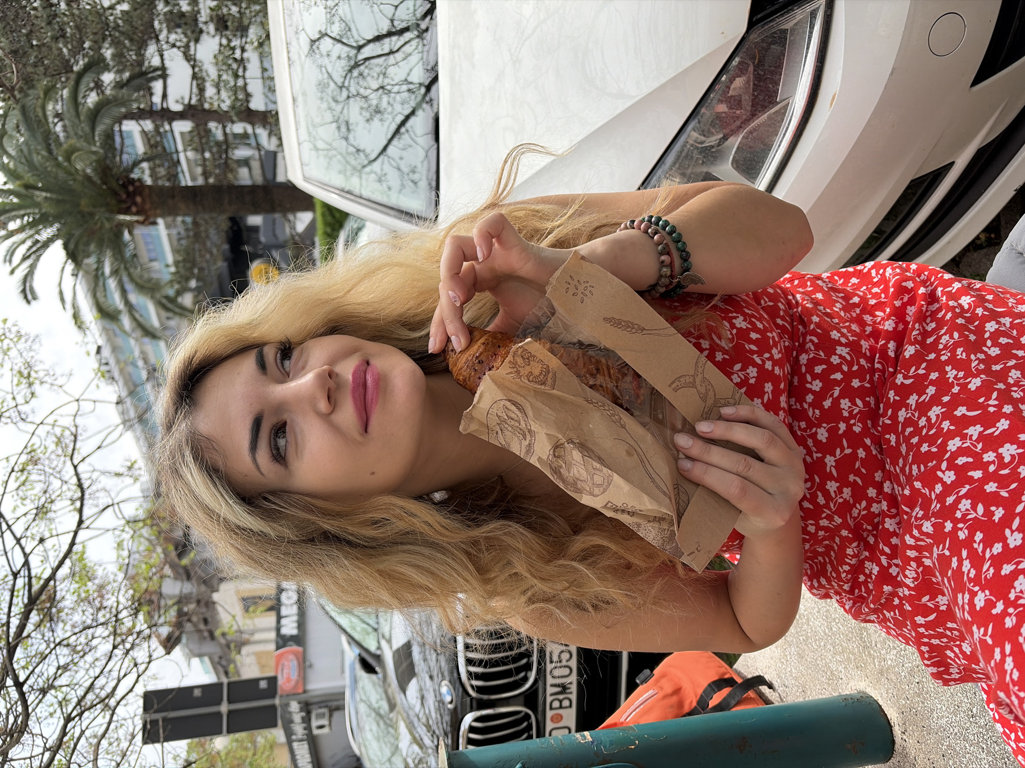 The image shows a woman standing in front of a white car, holding a bag of pastries. She has blonde hair and is wearing a red dress with a white floral pattern and bracelets on her wrist. She is holding a croissant in one hand and a brown paper bag containing two pastries in the other. The background includes trees, a palm tree, and a building with the word "MEGA" visible on a sign. In the lower part of the image, a black car with a license plate is partially visible. The location is likely in Budva, Montenegro, given the presence of a palm tree and urban setting.