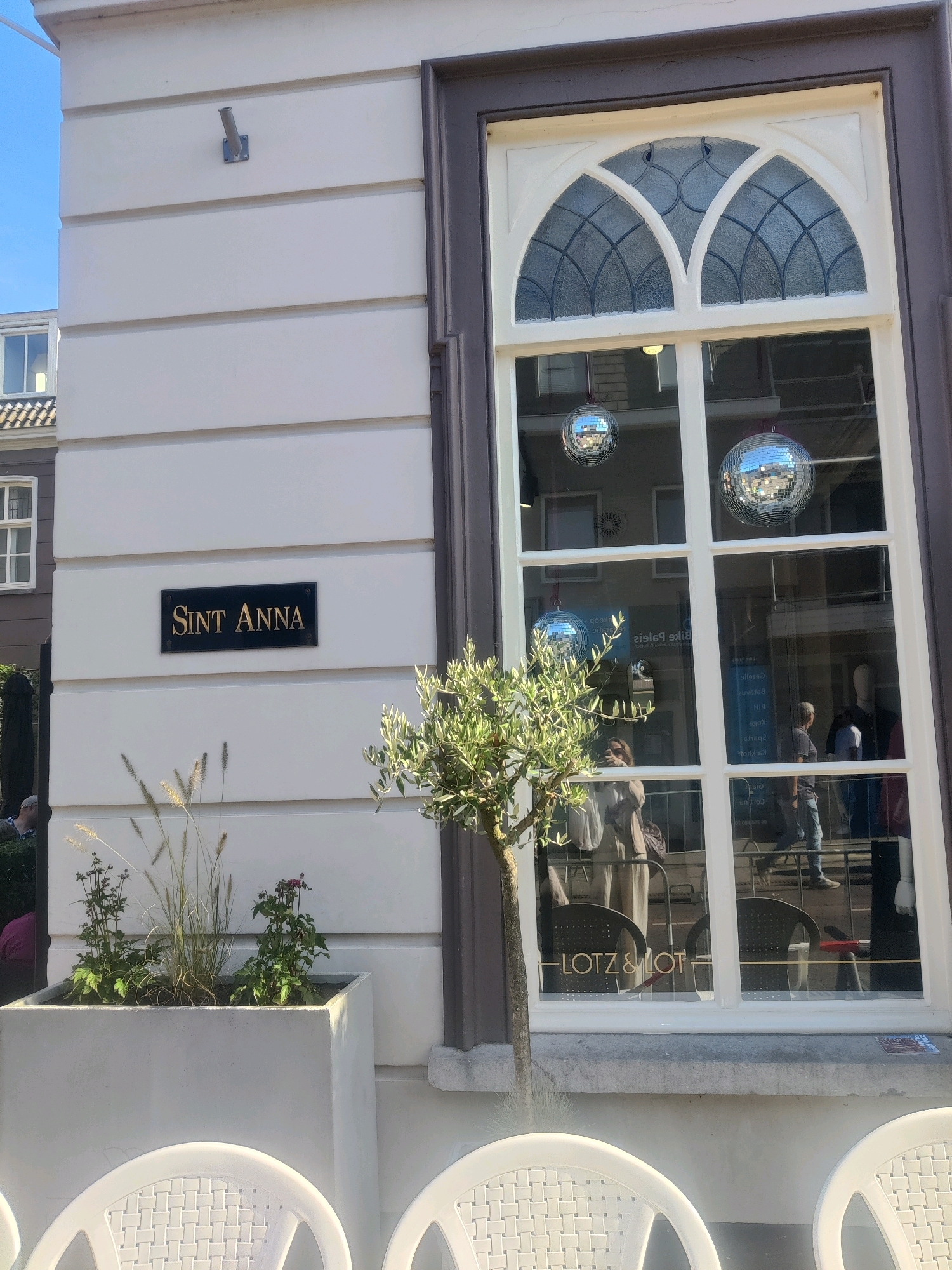 The exterior of a building is shown, featuring a cream-colored facade with horizontal panels and a dark-trimmed, arched window. A black sign with gold lettering reading "Sint Anna" is affixed to the facade. In front of the building, a large, light gray planter contains ornamental grasses and small plants. Three white chairs with woven seats are visible in the foreground. The window reflects the sky, building elements, and people inside what appears to be a shop or cafe. The reflection shows several disco balls hanging inside. The text "LOTZ & LOT" is visible on the window. The image is taken from a street-level perspective.