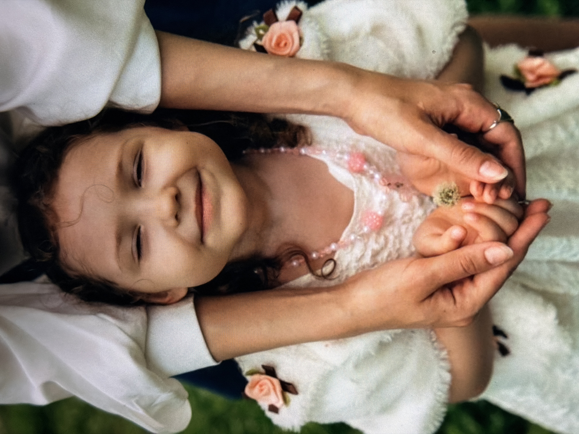 In a medium shot, a young girl with curly brown hair is centered in the frame, her eyes closed and a soft smile on her face. She wears a white, textured dress adorned with a pink beaded necklace and decorative floral accents on the shoulders. Her head is slightly tilted forward, and her hands are clasped together, holding a small dandelion. Adult hands are visible, cradling the girl's hands and body from behind, suggesting a protective embrace. The backdrop is a blurred expanse of green, indicating an outdoor setting. The lighting appears natural, casting gentle shadows. The overall tone is tender and familial.