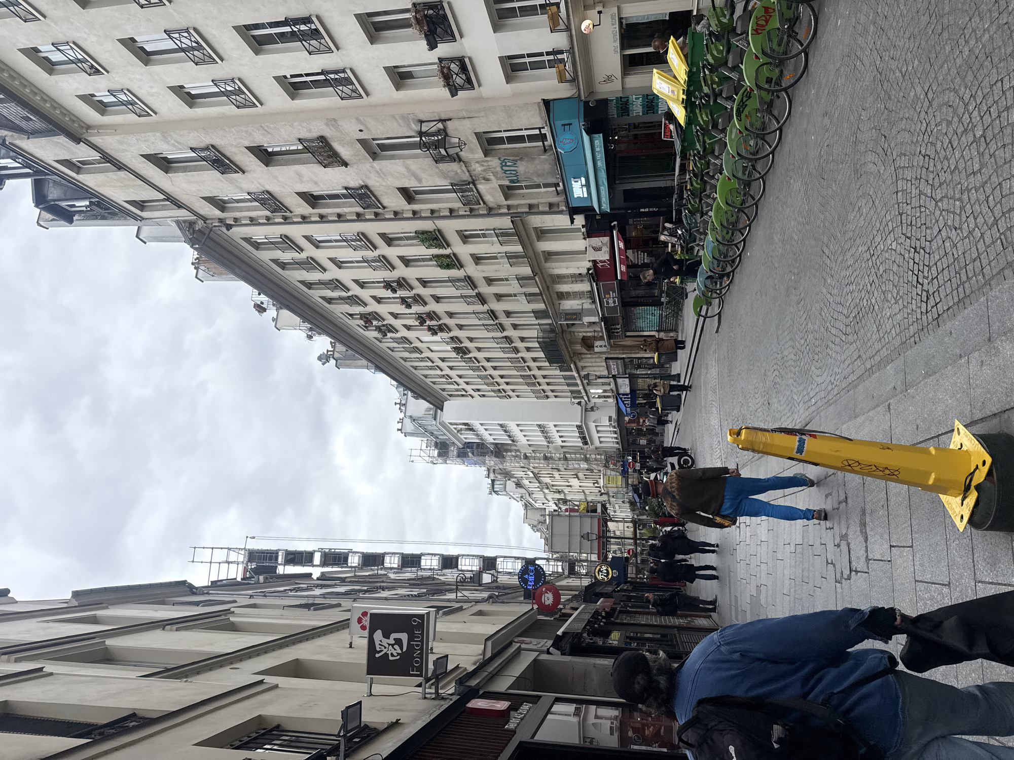 The image captures a street scene in Paris, France. The perspective is from the sidewalk looking down the street. Rows of buildings line the street, with numerous windows and architectural details visible. Several businesses are present, as evidenced by the signage. Bicycles are parked along the side of the street. People are walking on the sidewalk and in the street. A yellow traffic cone is present on the sidewalk. The sky is overcast. The signage includes "FONDUE 9," "ERIDGE," "Five," "TUPACT," and "100% FAIT MAISON".