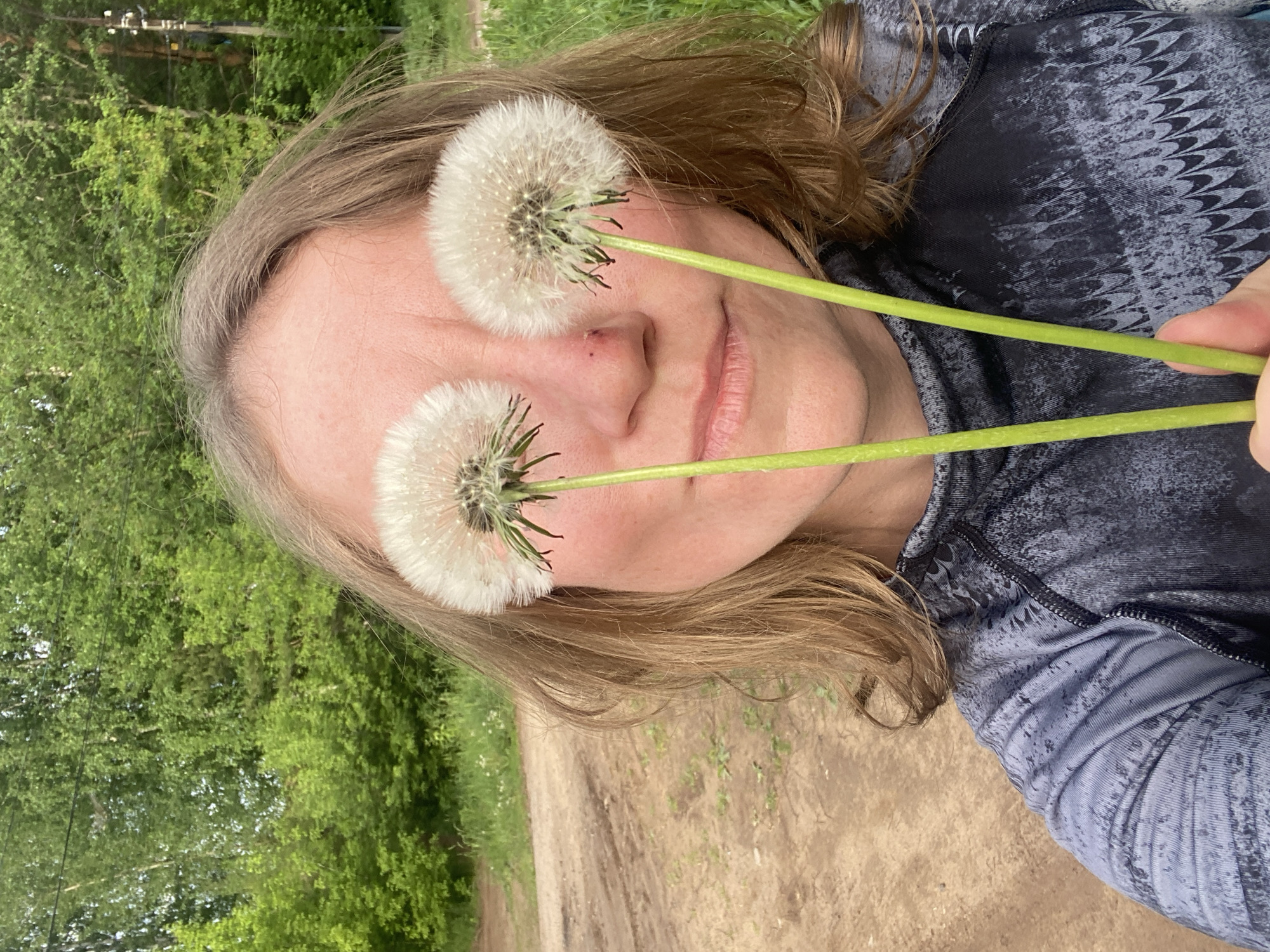 The image is a selfie of a fair-skinned woman with medium-length, light brown hair. Two dandelion seed heads are positioned to cover her eyes, with the green stems pointing downwards. She is wearing a gray patterned shirt. The background consists of green foliage and a dirt path. Based on the information provided, this location is in the city of Chaikovsky, Russia.