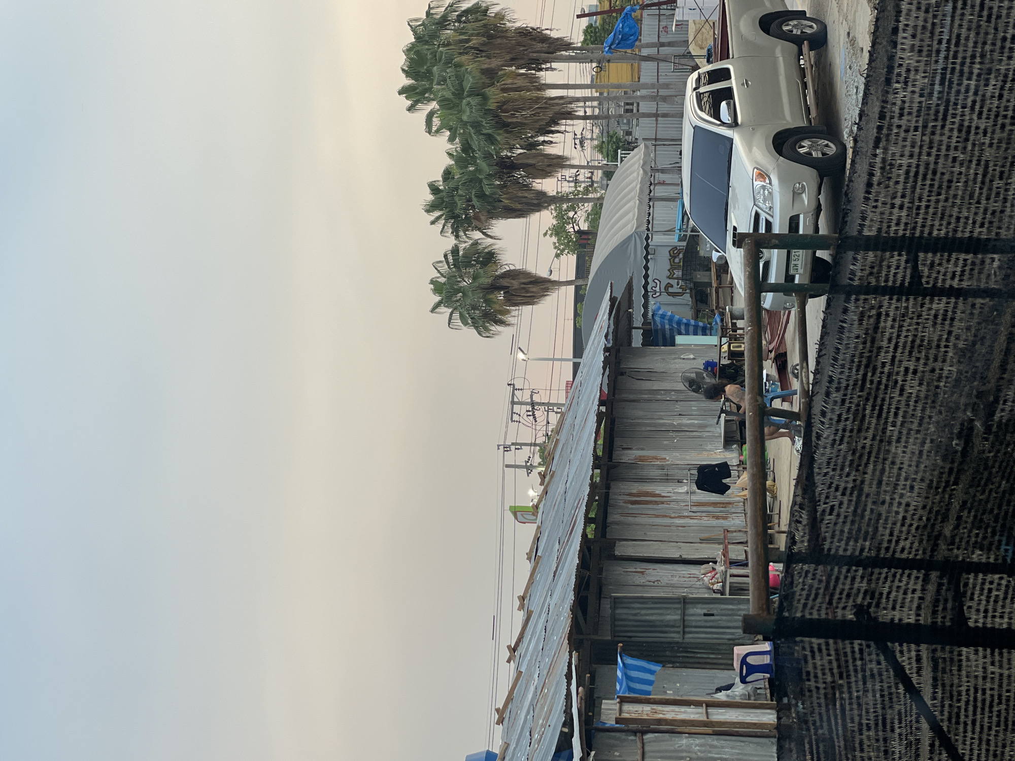 This street-level image captures an informal urban scene in Bangkok, Thailand, featuring rudimentary structures, tropical vegetation, and everyday activities under an overcast sky. The photograph is oriented vertically, but depicts elements typically arranged horizontally.
On the right side of the frame, a series of single-story structures constructed primarily from corrugated metal sheets serve as walls and roofs for what appear to be small shops, stalls, or dwellings. In the immediate foreground, a black woven mesh fence or screen partially obstructs the view. Under a canopy extending from one of the structures, a person with dark hair is seen from behind, sitting or crouching, engaged in an ordinary task. Nearby, a blue plastic chair, a black shirt hanging, and a fan are visible.
A light-colored pickup truck is parked on an unpaved road or shoulder alongside these structures. The truck's license plate, displaying Thai characters "นม 521", confirms the location as Thailand. Behind the truck, a white tent with a rounded roof stands.
To the left of the structures and road, a row of tall palm trees with green fronds lines the background, characteristic of a tropical climate. Utility poles and a network of power lines crisscross the upper middle ground, signifying an urban or semi-urban setting.
The sky is a hazy, diffused light grey, transitioning to a warmer, yellowish-beige hue on the far right. This soft, even lighting suggests an overcast day or perhaps early morning/late afternoon, creating a quiet and muted atmosphere. No strong shadows are present. Faint Thai script is visible on a signboard behind the truck but is illegible. The scene portrays a tranquil moment of daily life in a bustling yet informal part of the city.