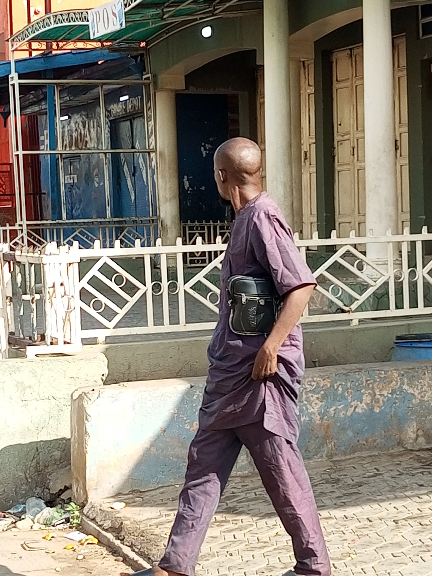 The image shows a bald man wearing a purple traditional outfit walking outdoors in front of a building with white metal fencing. The man is carrying a dark crossbody bag. Behind him, there are several large beige columns and cream-colored doors. Above the building entrance, a green awning with the word "POS" in white letters is visible. The ground is paved with light-colored interlocking bricks. To the left of the man, there is a low concrete structure with debris and some green vegetation. The scene appears to be a street in Arawa, Nigeria.