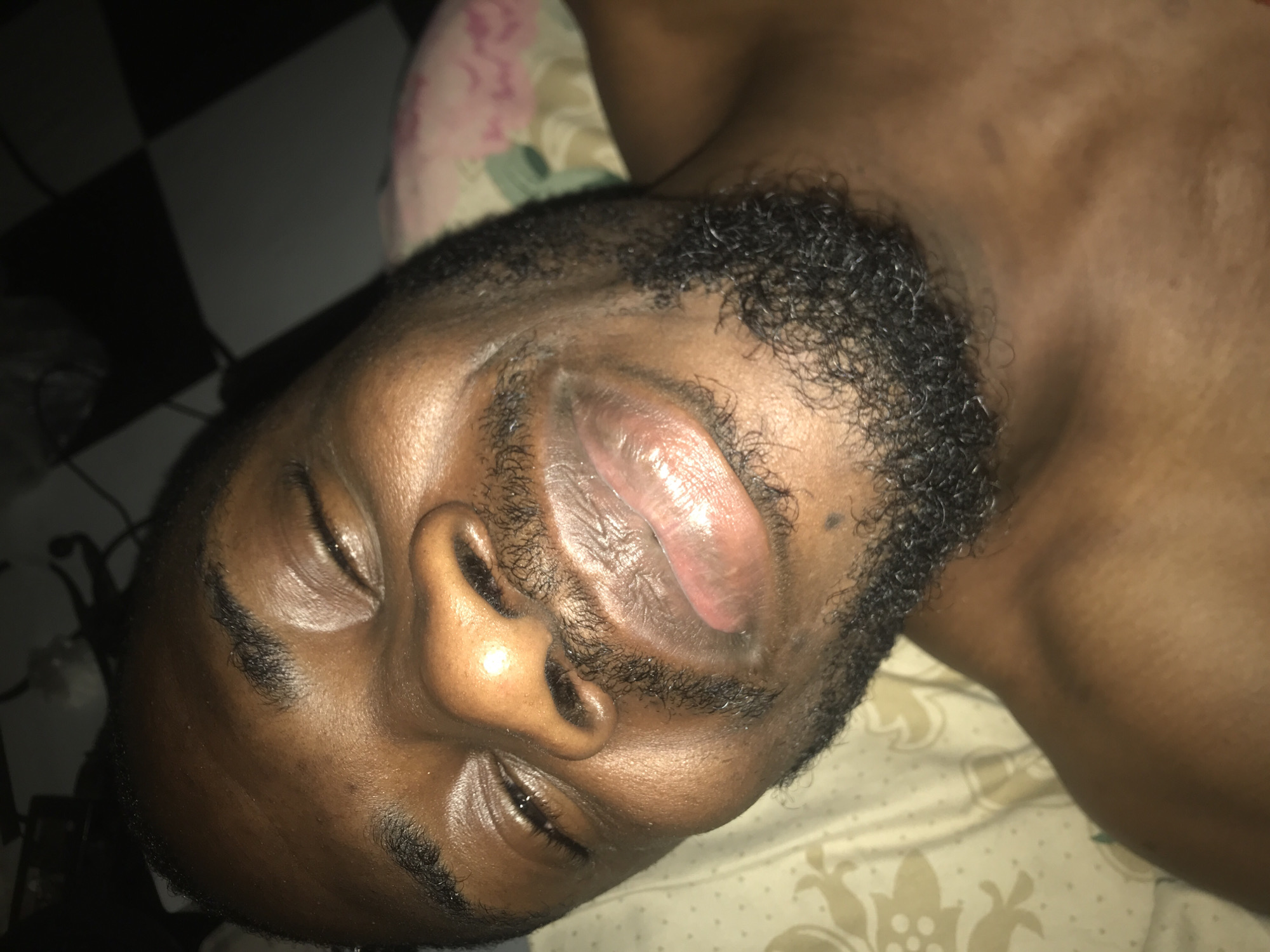The image is a close-up, downward-facing shot of a Black man's face, taken from a slightly elevated perspective. He appears to be lying down, with his head tilted back and his eyes partially closed. The lighting is dim, with a strong highlight on his forehead and nose, suggesting a light source from above and to the right.
The man has dark skin, a full beard and mustache, and short, dark hair. His lips are full and appear slightly moist. There is a faint pattern on the bedding behind him, which seems to be a light color with a floral or ornate design. To the right of the man's face, the background is dark, with a checkered pattern visible, possibly a floor.
The image does not provide enough visual information to determine the specific location within the city of Rafin Par, Nigeria.