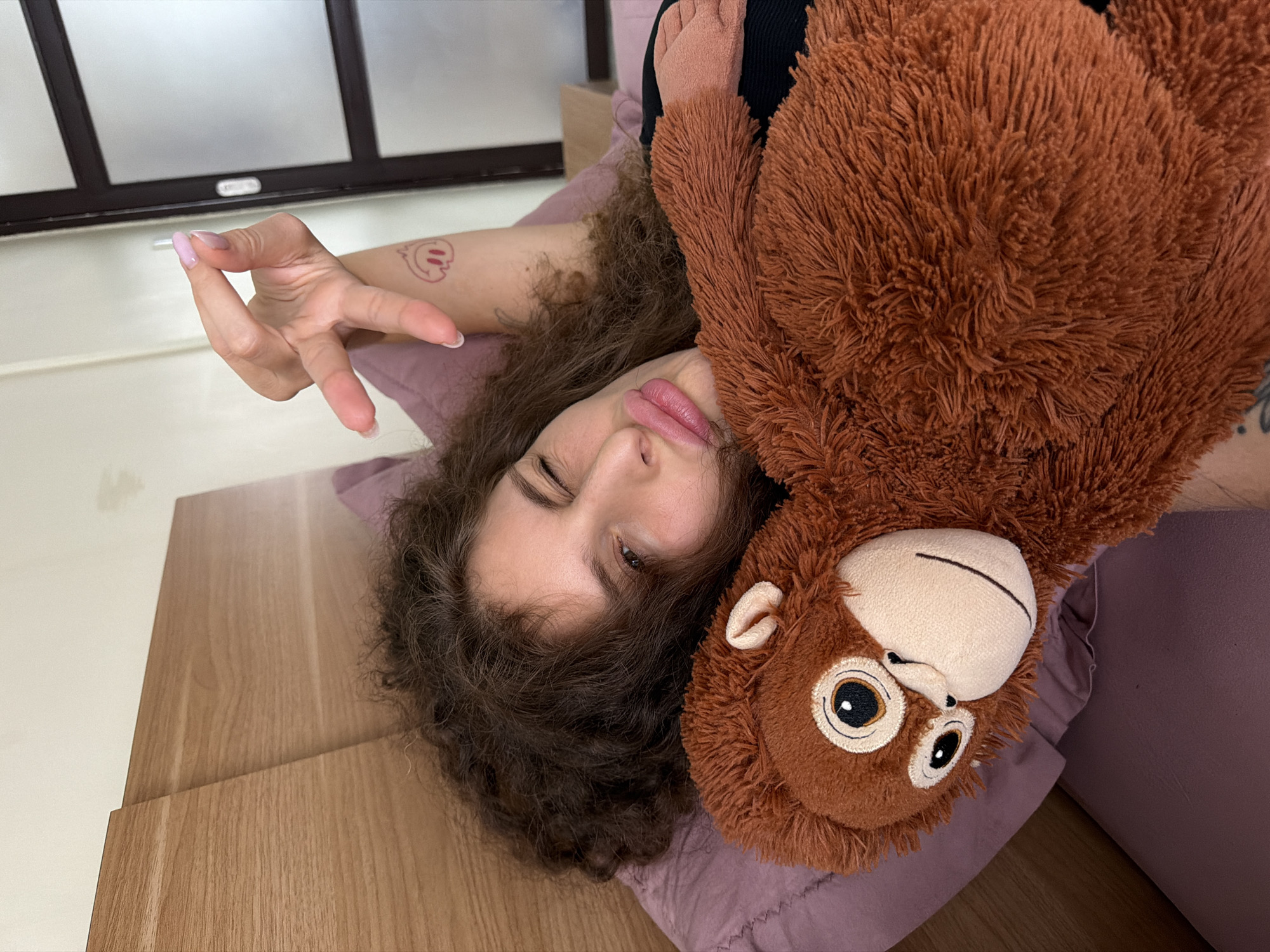 A woman with curly brown hair, lying in bed, holds a plush monkey toy. She is wearing a black top and has a tattoo on her left arm, which shows a smiley face. Her right hand is raised, and her nails are painted pink. The woman is wearing pink lipstick and her eyes are closed, with one eye squinting. The bed has a light brown wooden headboard and is covered with a purple duvet. In the background, a white wall and a sliding door with a frosted glass panel are visible. The image was taken indoors in a bedroom, likely in North Kuta, Indonesia.