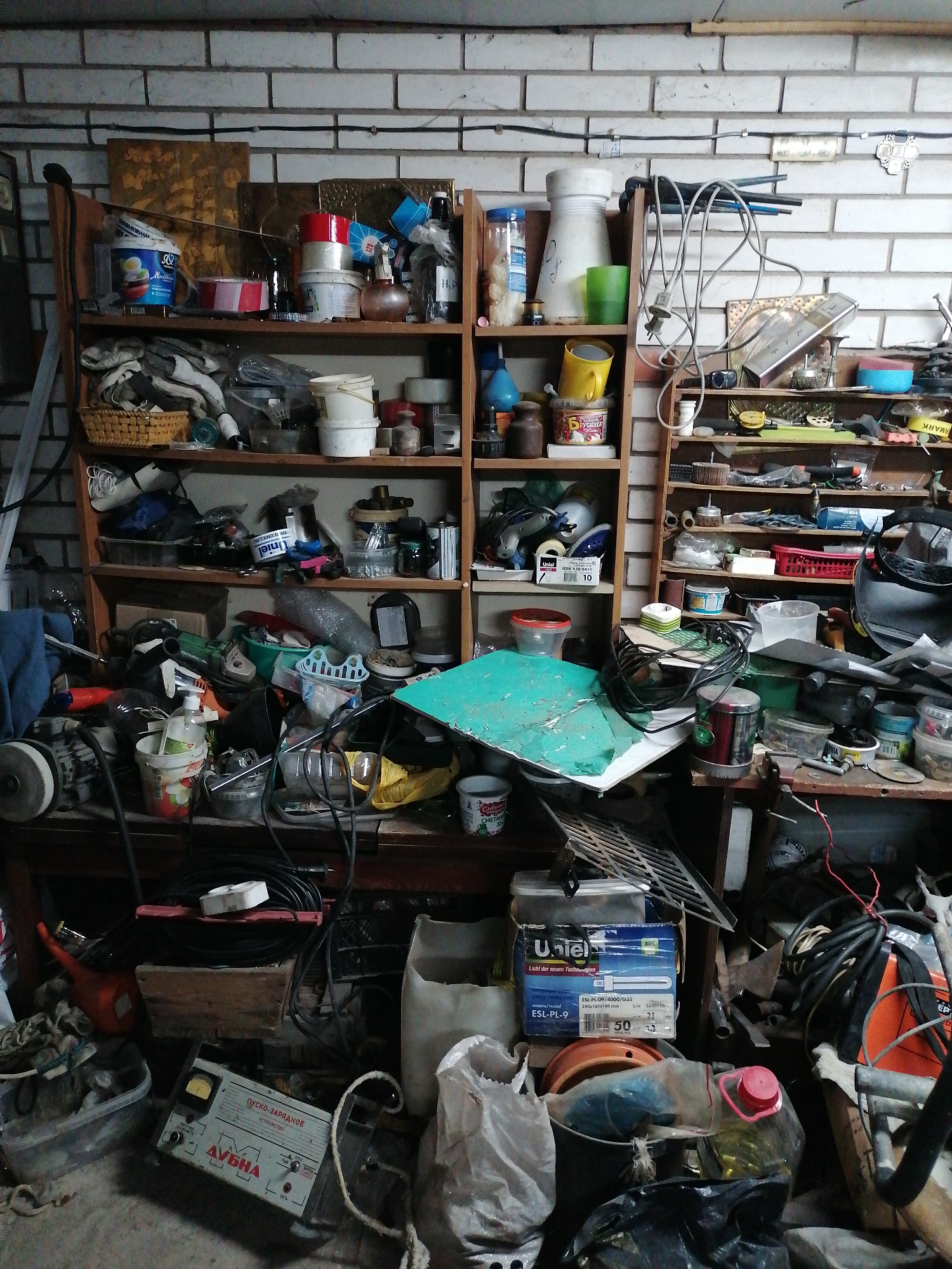 The image depicts a cluttered indoor space, likely a garage or workshop, characterized by a brick wall in the background. Two wooden shelves are filled with an assortment of tools, containers, and various objects, suggesting a storage area. A workbench beneath the shelves is covered in more tools, equipment, and miscellaneous items. The floor area is also littered with objects, including power tools, boxes, and bags, indicative of a workspace in disarray. A device labeled "ДУБНА" is visible on the floor, as well as a box labeled "Uniel."