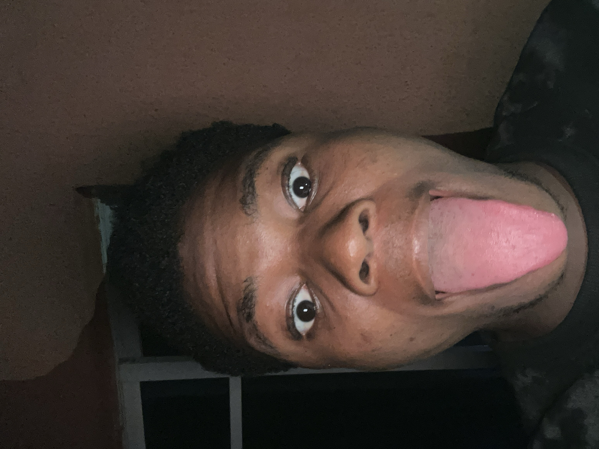 A close-up, eye-level, indoor selfie shows a young Black person with dark, curly hair and brown eyes sticking out their tongue. The person is looking directly at the camera with wide eyes and a slightly open mouth. Their tongue is extended, and it is bright pink. They are wearing a black t-shirt. The background is dimly lit and appears to be a wall with a window or doorway on the left side. The overall lighting suggests the photo was taken at night or in a dimly lit room. The specific location in Ihiagwa, Nigeria, is not discernable from the image itself.