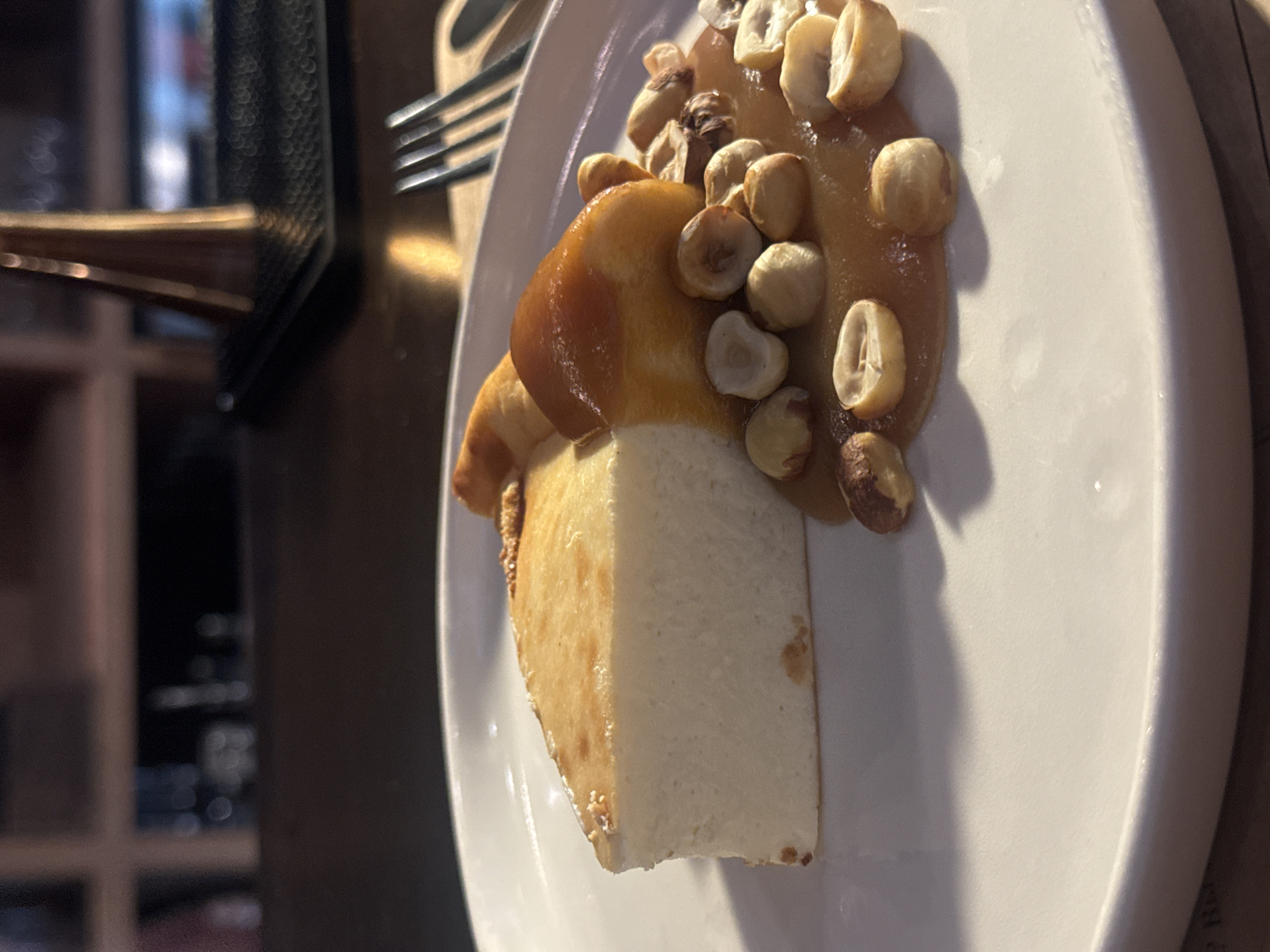 A close-up, eye-level shot shows a slice of cheesecake on a white plate. The cheesecake is a thick wedge with a smooth, off-white interior and a lightly browned, slightly puffed crust. It is topped with a swirl of glossy, amber-colored sauce and a scattering of whole and halved roasted hazelnuts. A fork is visible to the right of the plate, with its tines resting on the rim. The background is blurred, suggesting an indoor setting with warm, dim lighting. A wooden surface and shelves with out-of-focus objects are visible. The image does not contain any specific visual cues to pinpoint its location in Krasnoyarsk, Russia.