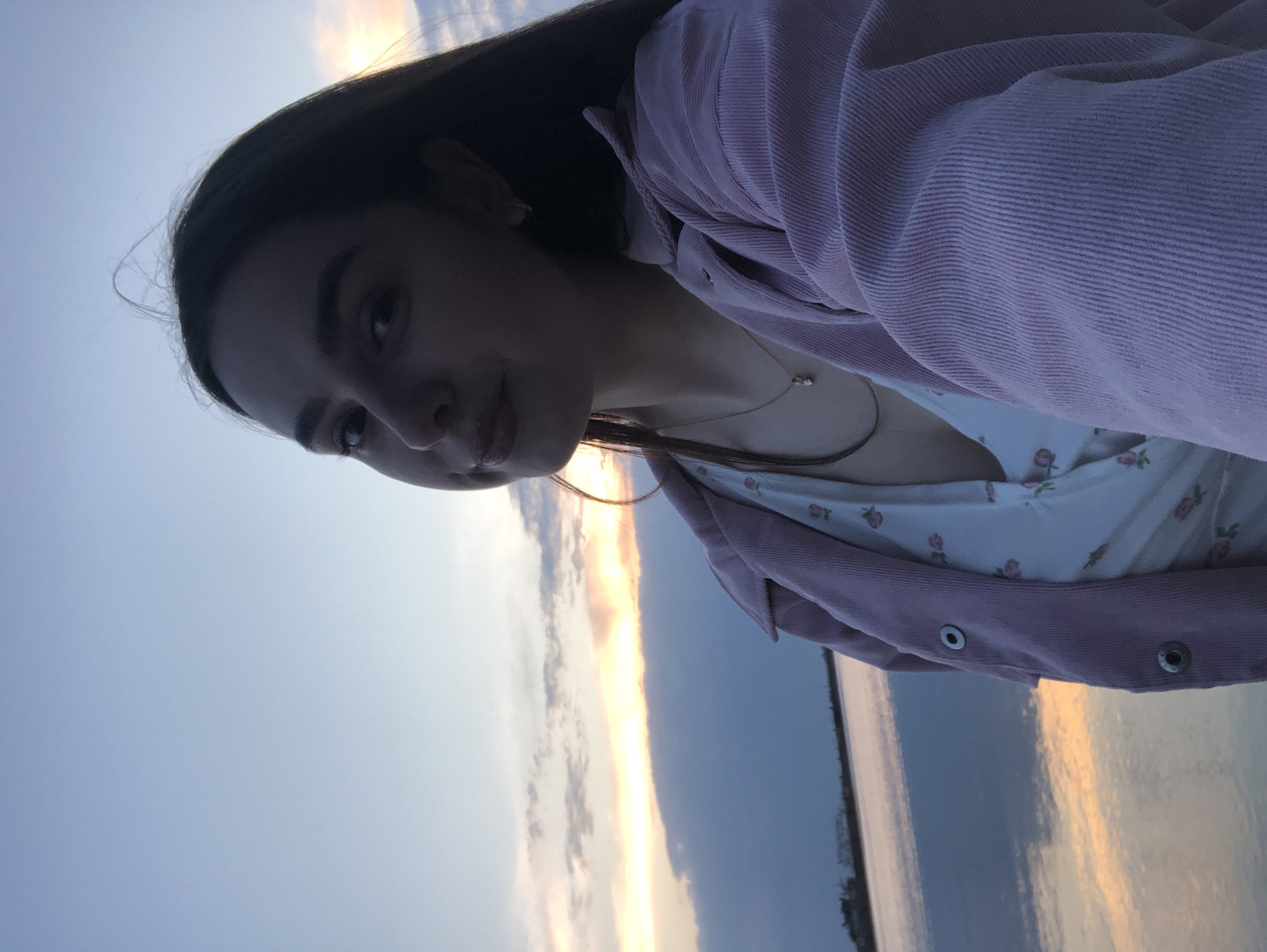 The image is a selfie taken outdoors, likely near a body of water at dusk or dawn. A young woman is in the foreground, wearing a white floral print top and a pink corduroy jacket. She is slightly angled to the left of the frame, looking at the camera.
The background shows a sky with a mix of blue and orange hues, suggesting either sunrise or sunset. There is a body of water, possibly a river or lake, reflecting the colors of the sky. The location is presumed to be in Chaikovsky, Russia, though there are no distinct landmarks or signage visible in the image to confirm this.