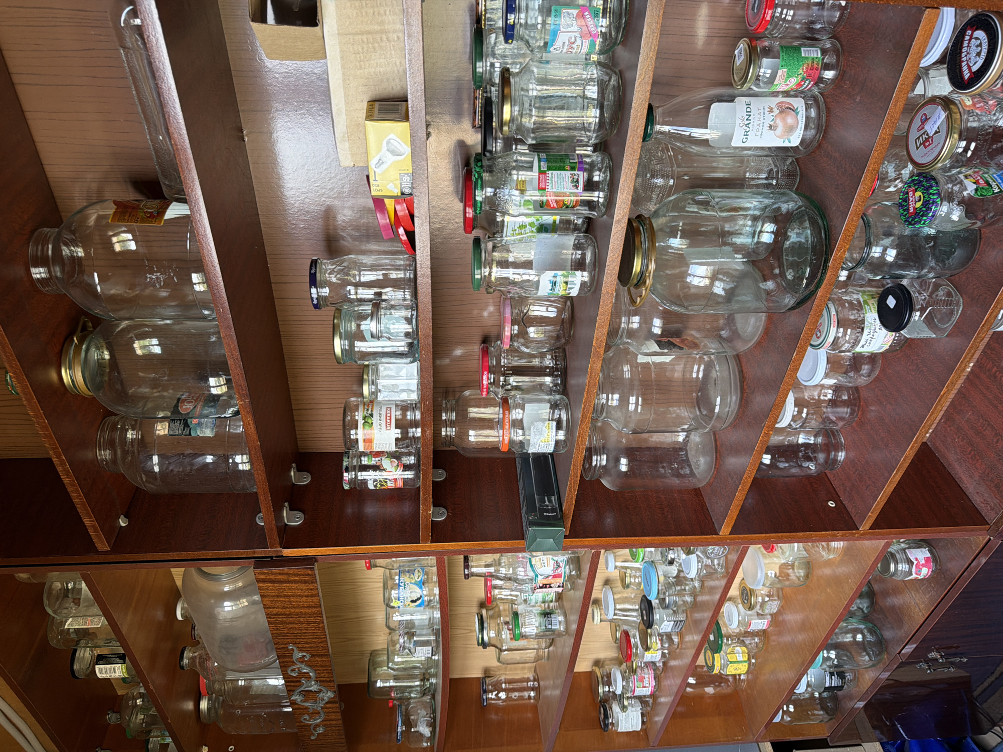 The image depicts a wooden cabinet filled with various glass jars of different sizes and shapes. Some jars contain labels with Russian text, suggesting the location might be in Ягул, Russia. The jars are organized on shelves within the cabinet. A few jars have lids on them, while others are empty. The cabinet appears to be indoors.