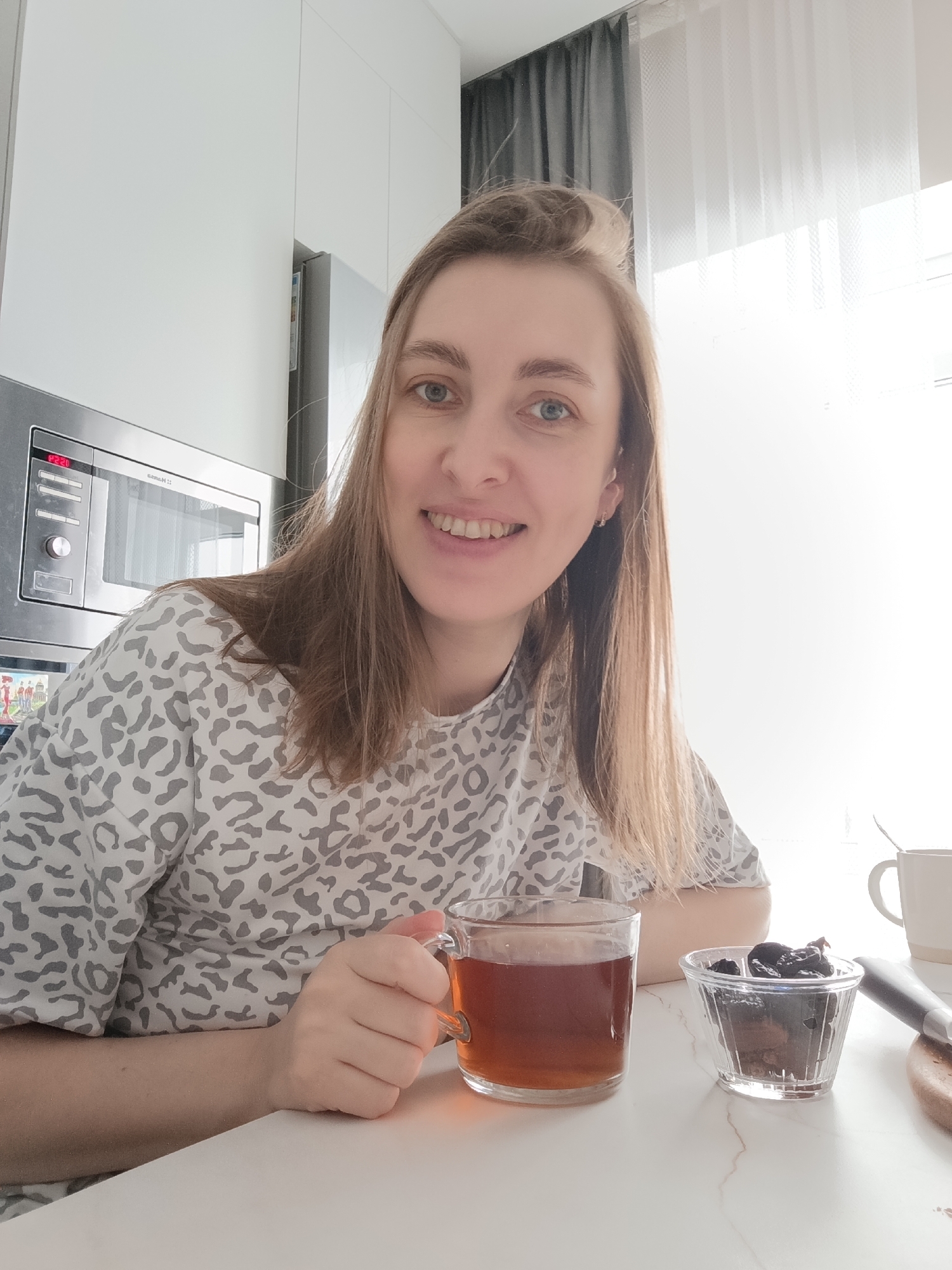 A female subject with light-brown hair and blue eyes is seated at a white, marble-patterned table, smiling directly at the viewer. She is wearing a white and gray leopard-print long-sleeved shirt. Her right hand holds a clear glass cup containing a dark beverage. On the table, to her right, is a small clear glass bowl filled with dark dried fruit, likely prunes. Partially visible further right are a white ceramic mug with a spoon and a wooden cutting board with a dark-handled utensil.
The background reveals a modern kitchen or dining area. To the subject's left, white glossy cabinetry is visible, along with a built-in stainless steel microwave or oven unit displaying "12:20" in red digits. A portion of a silver refrigerator and a small colorful item are also present. To the subject's right, a brightly lit window with white sheer curtains and dark gray curtains pulled to the side is visible. The scene is situated in a residential interior in Chaikovsky, Russia.