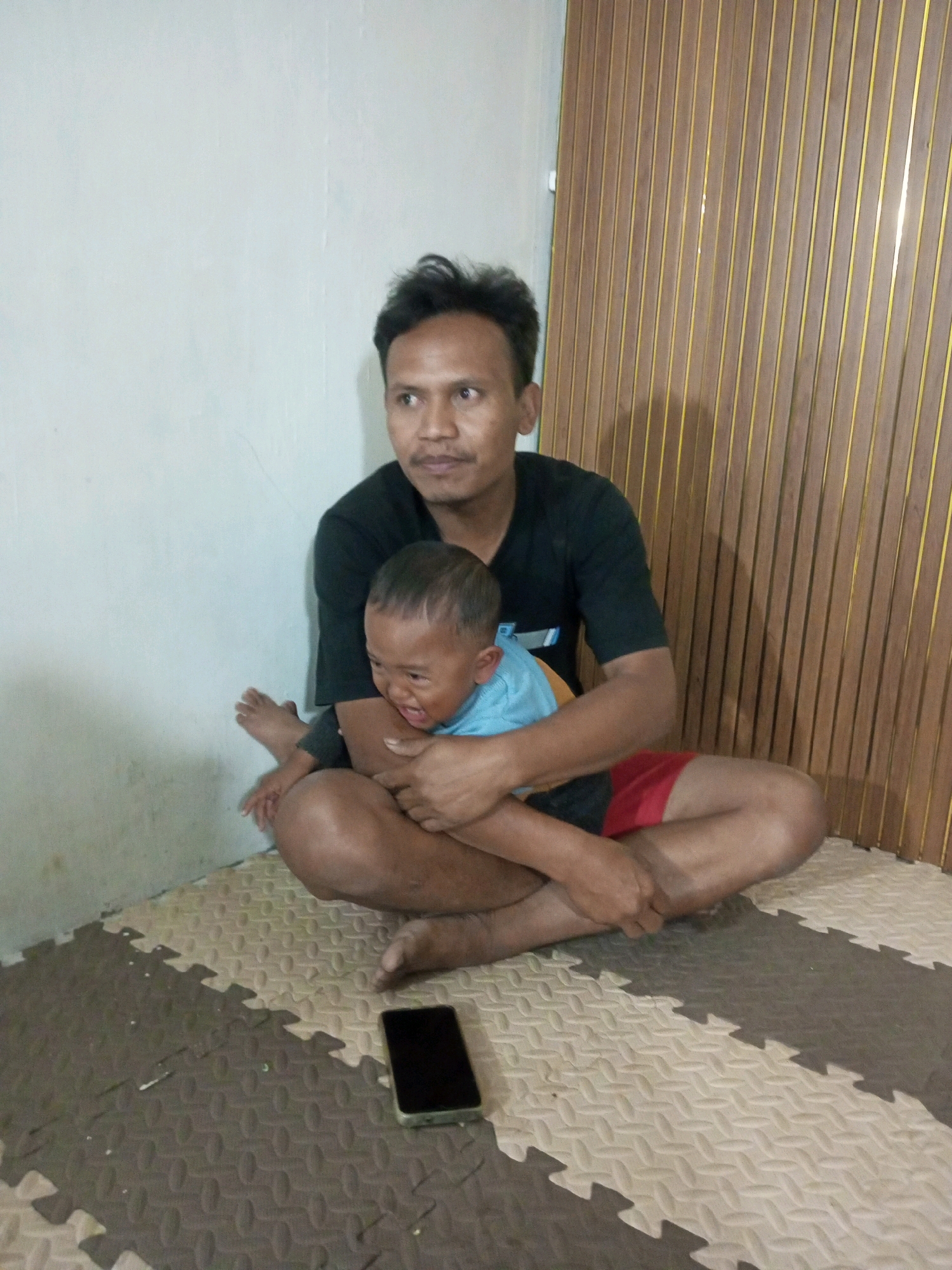 The image shows a man sitting cross-legged on a mat, holding a young child. The man is wearing a black t-shirt and red shorts. The child is wearing a blue shirt and appears to be crying. They are positioned in front of a wall with vertical wooden panels. A smartphone is visible on the mat in the foreground. The environment appears to be indoors, likely a home. The photo captures a moment of interaction between the man and child.