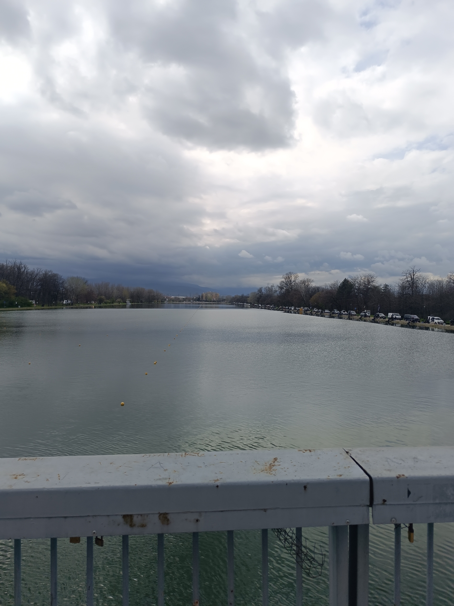 This image depicts a view of the Rowing Canal in Plovdiv, Bulgaria. The perspective is from a bridge overlooking the canal. The water surface is relatively calm with visible ripples. A row of yellow buoys is visible, extending into the distance, likely marking a lane for rowing. The banks of the canal are lined with trees, some of which appear to be bare or sparsely leaved. On the right bank, a line of parked vehicles is present. In the background, there are buildings visible against a cloudy sky with overcast conditions. The bridge's railing is in the foreground, featuring a grey color. There are small locks attached to the railing in the foreground.