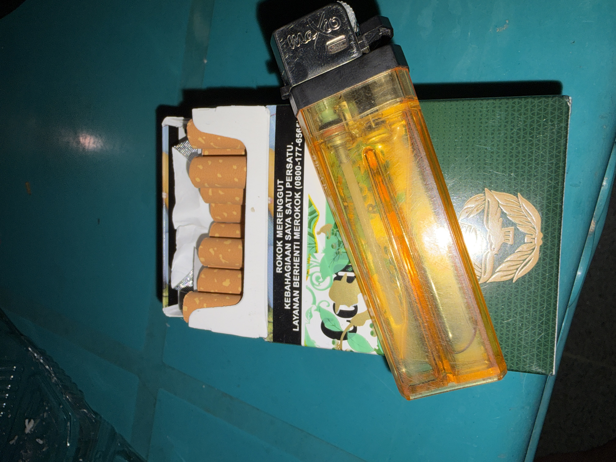 The image shows a close-up, high-angle shot of a pack of cigarettes and a transparent orange lighter placed on a green surface. The cigarettes are visible inside the open pack, with their orange filters facing upwards. The lighter is positioned diagonally across the cigarette pack. The green surface appears to be a table or a similar flat object with a subtle textured pattern. The background is a teal-colored surface, possibly a wall or another table. The lighting in the scene is dim, creating shadows and highlighting the objects. The overall impression is that of a still life composition. The provided information about the location in Tupah, Indonesia is not visually supported by the image content.