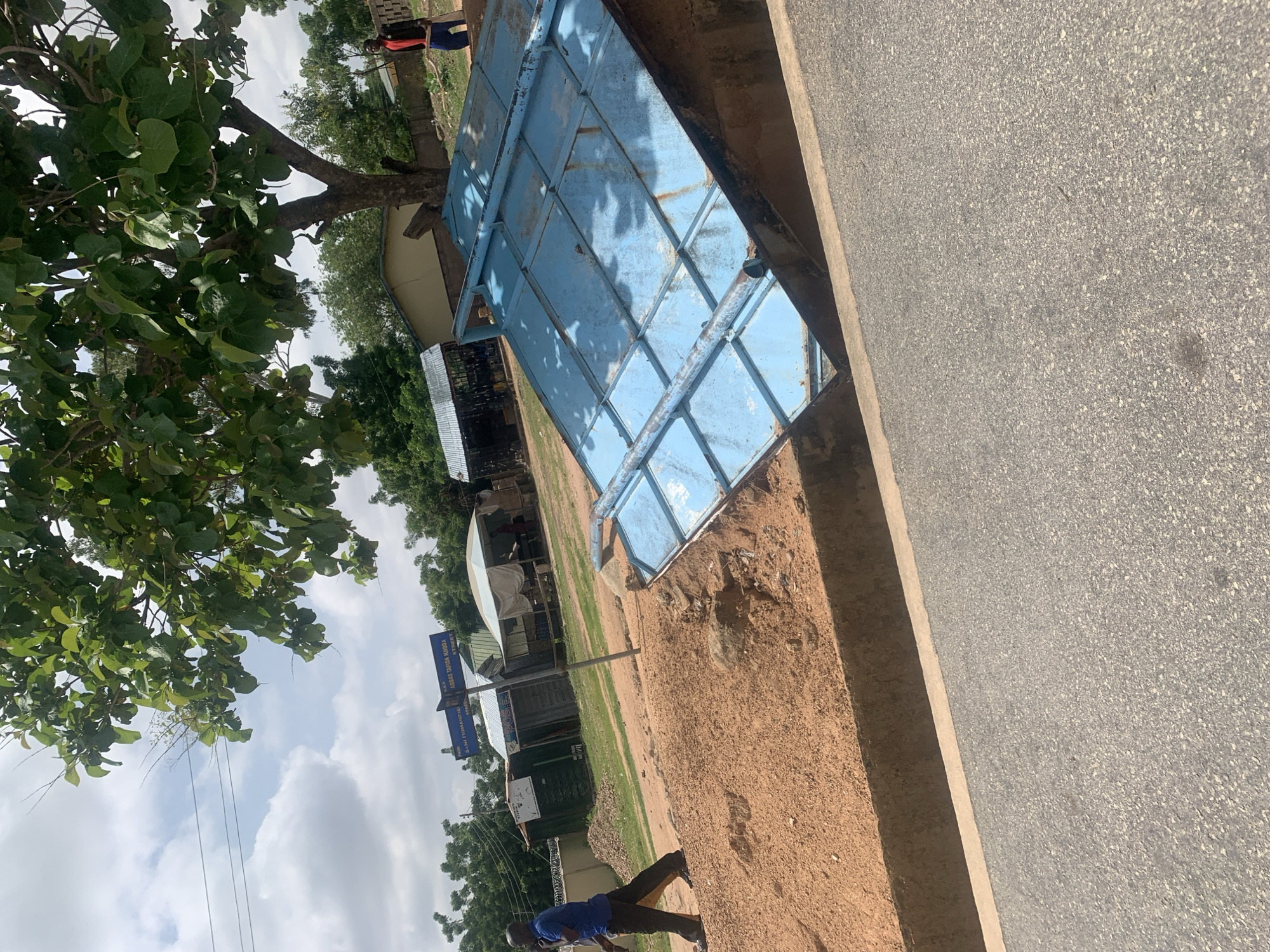 A man in a blue shirt and dark pants walks to the left on a dirt path. He is in the foreground, to the left of a large, tilted, blue metal structure that appears to be a gate or part of a fence. The structure is resting in a ditch. Behind the man and the structure are several small buildings with corrugated metal roofs and awnings. A street signpost with multiple signs is visible in the background. Trees with green leaves are in the background and overhead. A second person, wearing a red shirt and dark pants, stands to the right, further in the background, holding a blue object. The scene appears to be an outdoor environment in Jalingo, Nigeria, with a road visible in the bottom right of the frame.