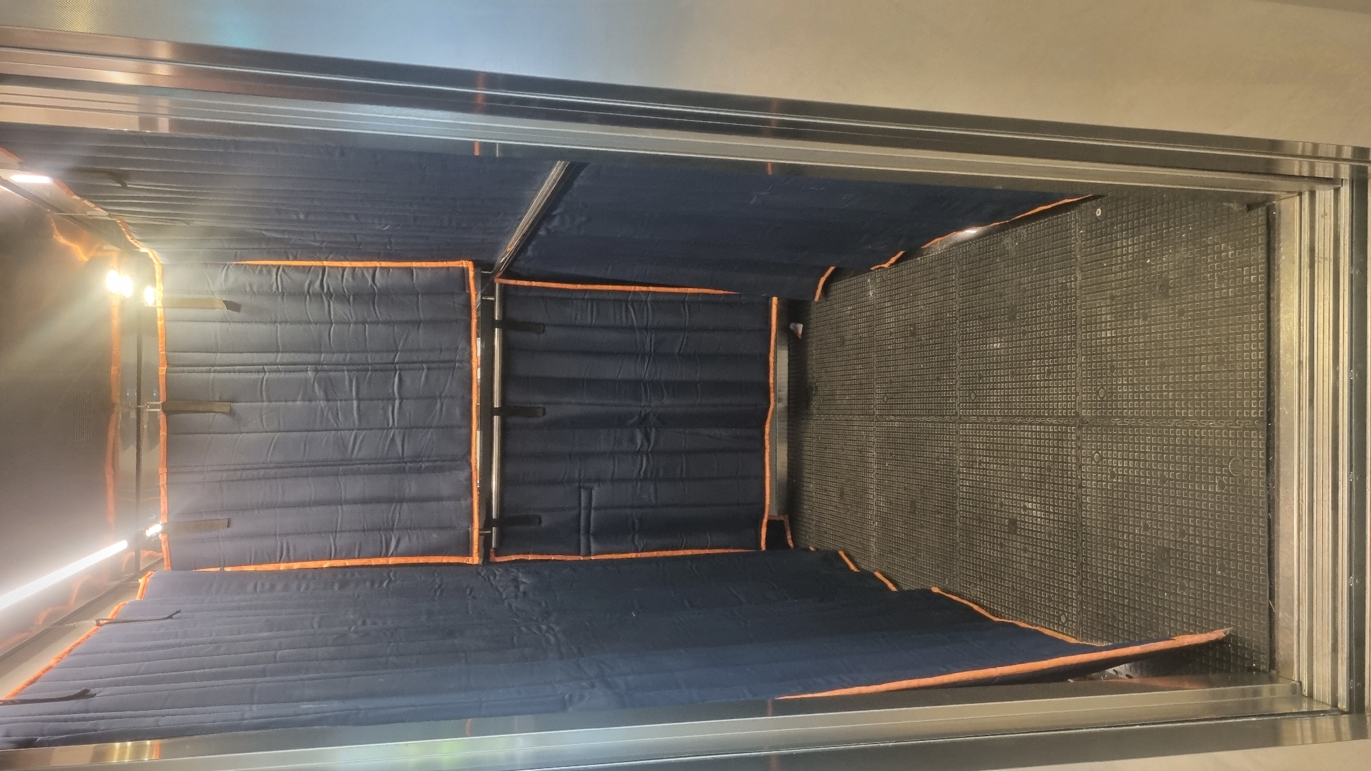 An interior view of an elevator shaft shows dark blue padded curtains with orange trim lining the walls, suspended from overhead metal bars. The curtains are segmented and appear to be made of a quilted fabric. The floor of the elevator is covered with a textured grey mat. Overhead, two bright LED lights illuminate the space. The right wall of the elevator shaft is a polished metal surface. The scene is shot from a slightly low angle, looking into the shaft.