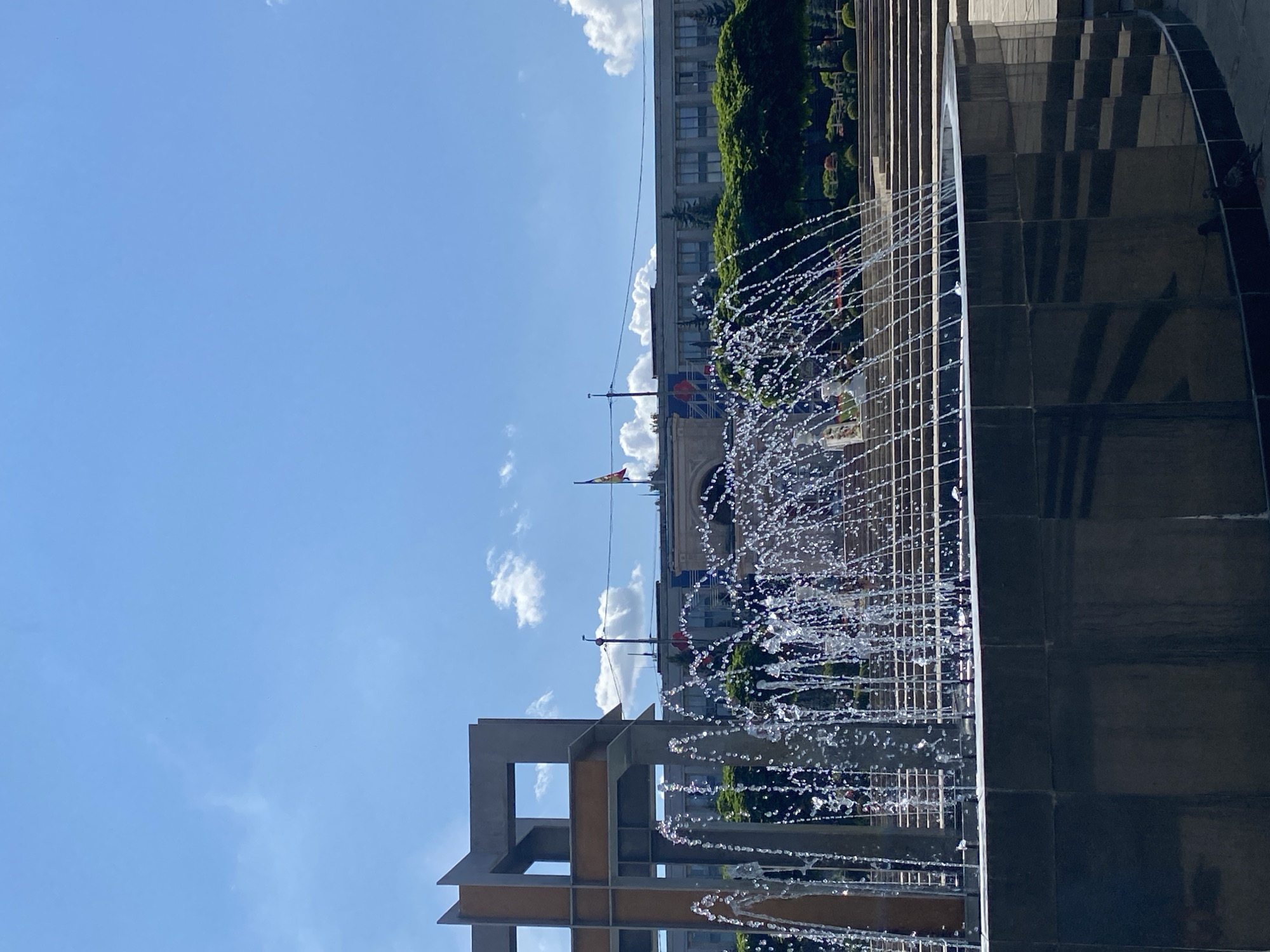 This is a photograph of the Great National Assembly Square in Chișinău, Moldova, on a sunny day. In the foreground, a modern fountain sprays water upwards, creating a shimmering curtain of droplets. The fountain's base is made of dark, reflective material. Behind the fountain, a large, abstract sculpture made of concrete and metal is visible. Further back, a grand building, likely the Parliament or Presidential Palace, stands prominently with flags flying in front. Lush green trees and a terraced staircase are also part of the scene, suggesting a well-maintained public space. The sky is a vibrant blue with scattered white clouds. Two pigeons are visible near the base of the fountain in the lower right corner of the image.