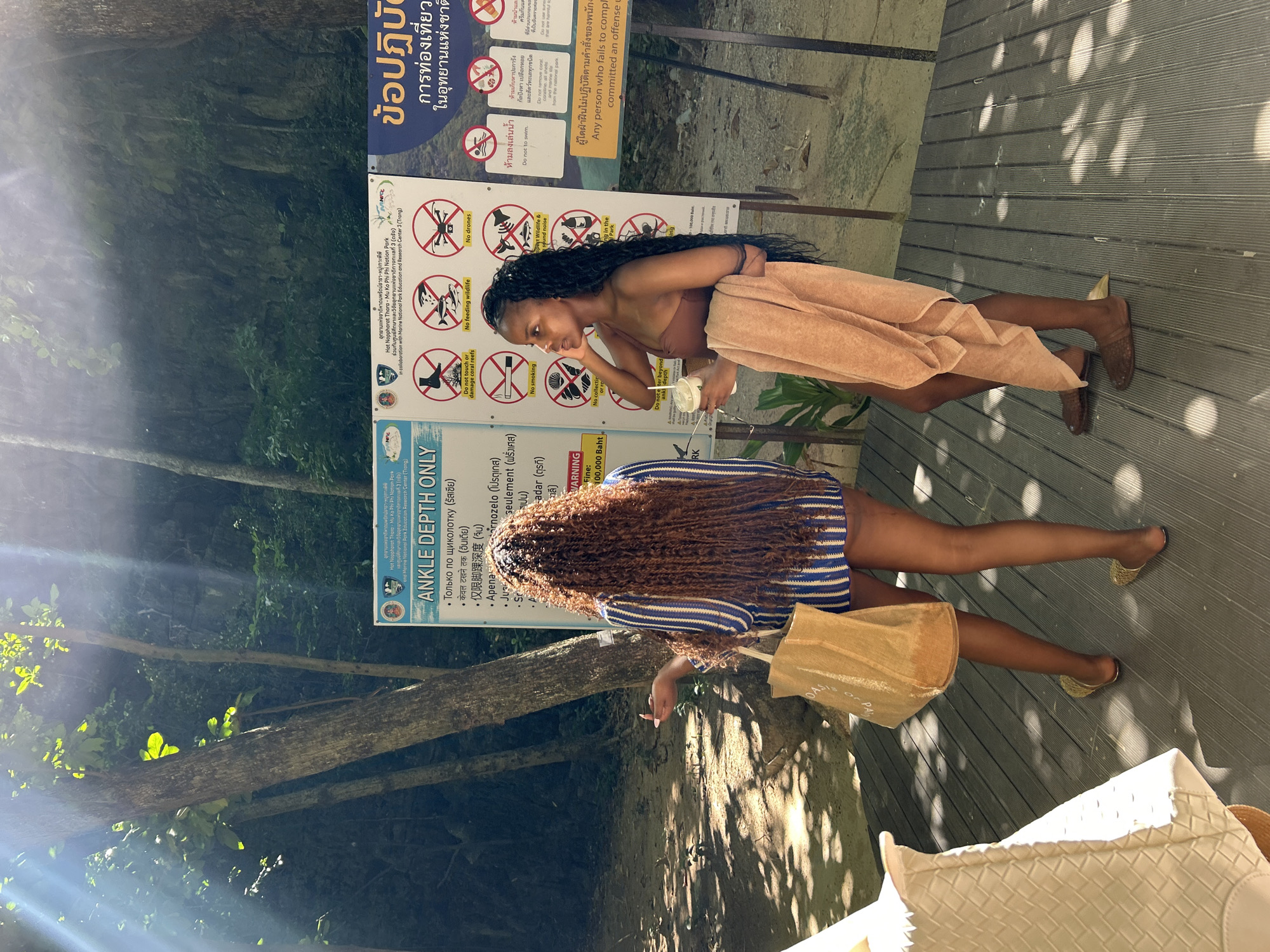Two young women are standing on a wooden boardwalk in Ao Nang, Thailand, near a sign with park rules in multiple languages. The woman on the left, with long, curly brown hair, wears a blue and white striped shirt and is holding a light brown tote bag. The woman on the right, with long dark curly hair, wears a brown bikini top and has an orange towel draped over her arm. She is holding a cup in her right hand. Behind them, a rocky cliff face is covered in lush green foliage, and sunlight filters through the trees, casting dappled shadows on the boardwalk. The signpost behind them contains regulations for visiting a national park, including prohibitions against touching coral reefs, feeding wildlife, and using drones.