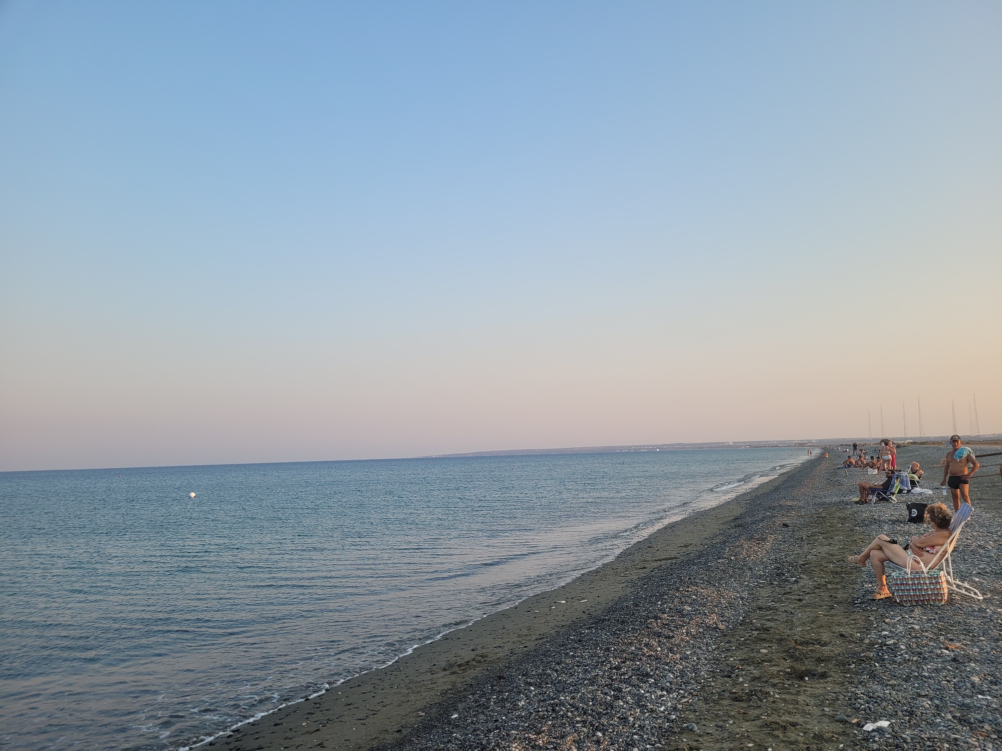 An outdoor, medium-shot image displays a wide, pebble-strewn beach in the Sovereign Base Area of Akrotiri and Dhekelia, with the sea on the left and a pale sky on the right. The water is calm and a light blue, with a white buoy visible in the distance. The beach is populated by several individuals relaxing on chairs or walking along the shoreline. In the foreground, a woman sits in a striped beach chair, facing away from the camera. Further down the beach, a man stands with a towel draped over his shoulder, looking towards the sea. Other people are scattered along the beach, some seated, others standing, and some walking closer to the water. The background shows a hazy line of land and structures, likely part of the Sovereign Base Area. The overall scene depicts a peaceful, late afternoon at the beach.
