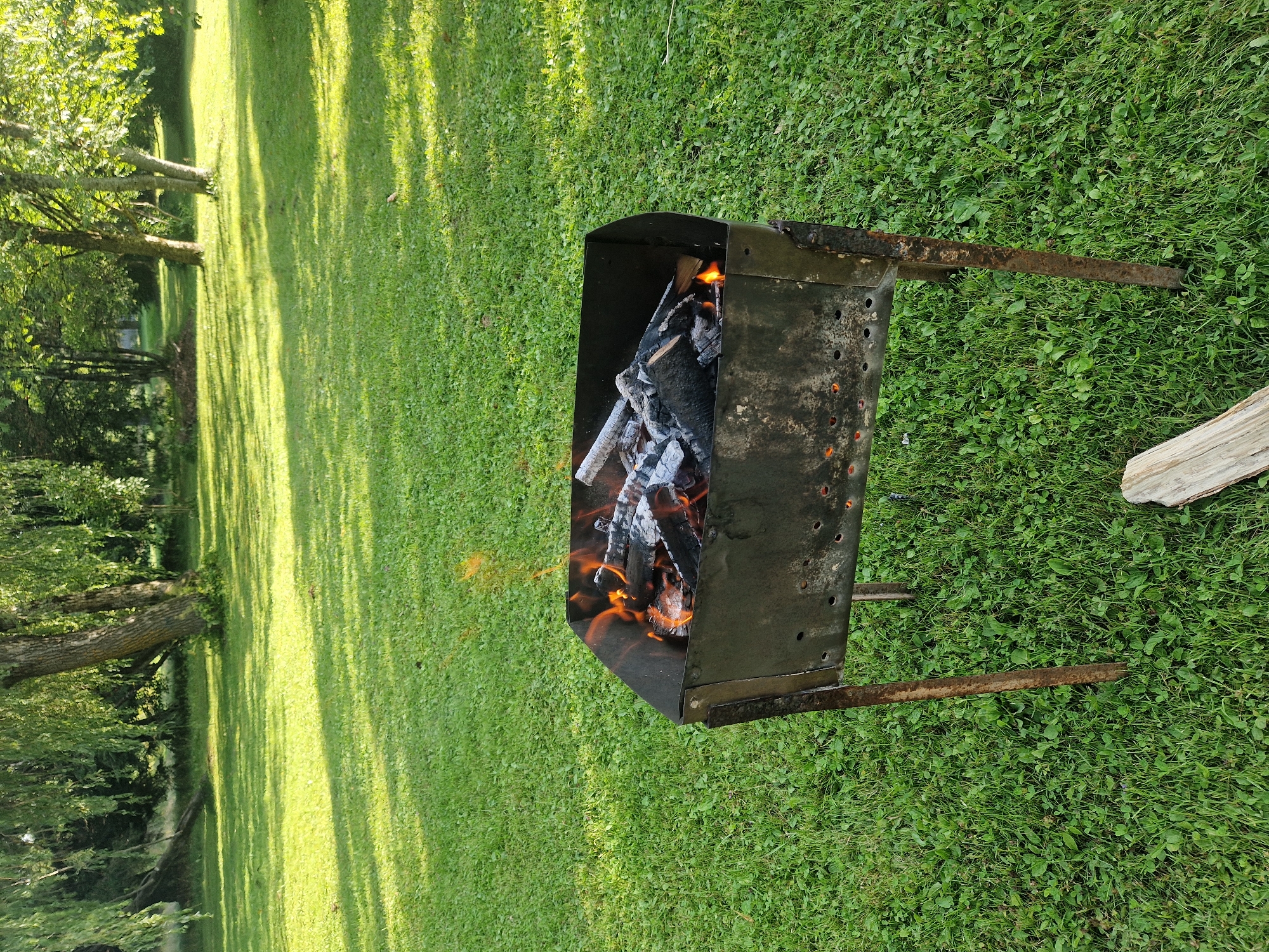 In the city of Zemkopības institūts, Latvia, a metal barbecue grill with burning wood sits on a lush green lawn. The grill, which appears aged with some rust and small holes along its sides, is filled with logs and embers, with visible flames licking upwards. The surrounding area is a well-maintained grassy field, bathed in dappled sunlight filtering through the leaves of mature trees. These trees, with their full canopies, form a natural backdrop to the scene, suggesting a park or garden setting. A piece of light-colored wood lies on the grass in the foreground, to the right of the grill. The overall impression is one of a calm, outdoor environment, possibly prepared for a gathering or event.