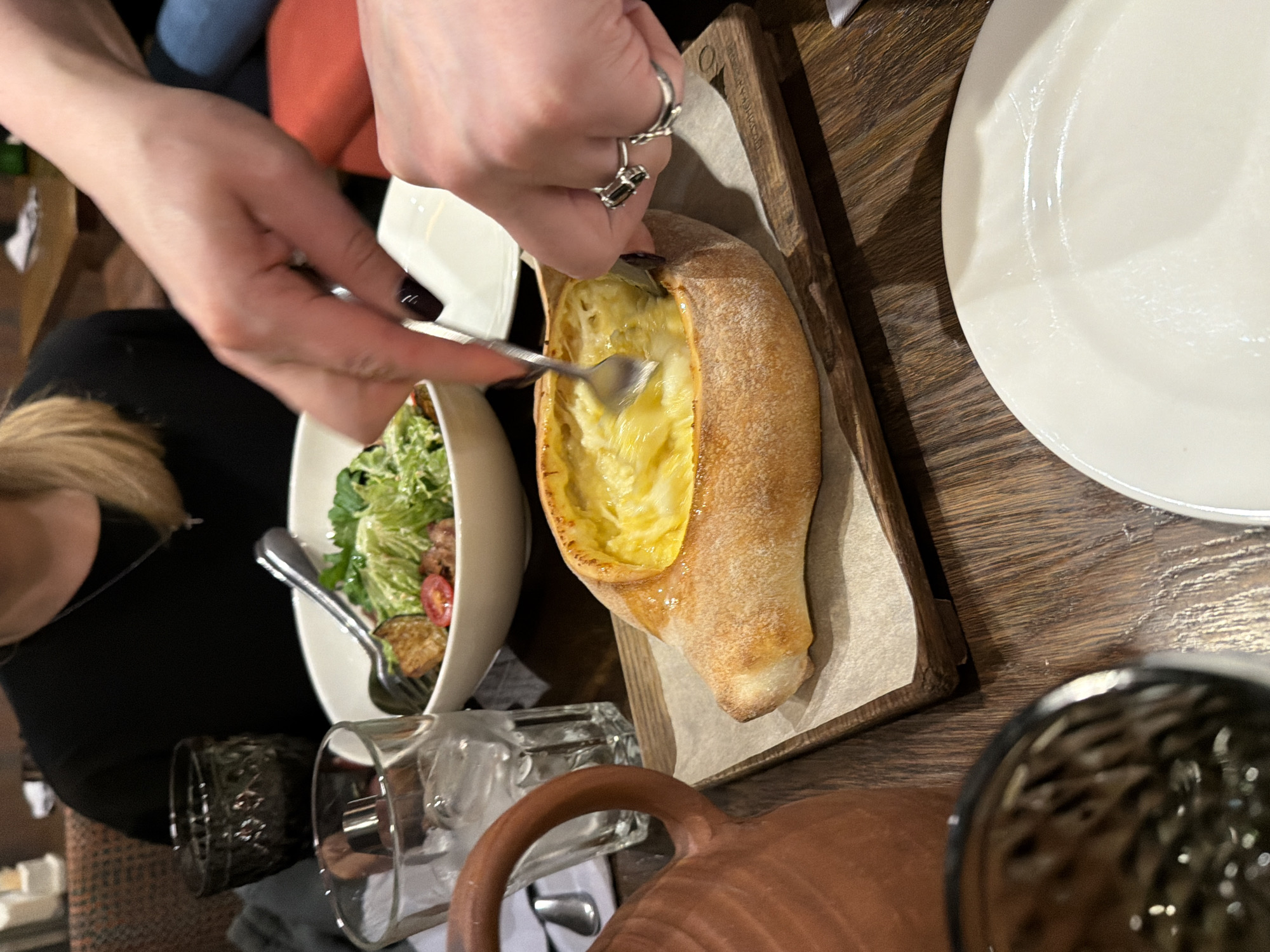 The image shows a table setting, with a focus on a person using a fork to interact with a Khachapuri Adjaruli, a Georgian cheese bread, served on a wooden board. To the left, there's a salad in a white bowl. There are also water glasses and a ceramic pot on the table. A white plate is situated to the right of the Khachapuri. The scene likely takes place in a restaurant. While the image does not provide direct evidence, the presence of Georgian cuisine might suggest a restaurant serving that type of food in Saint Petersburg, Russia, if the location is assumed.