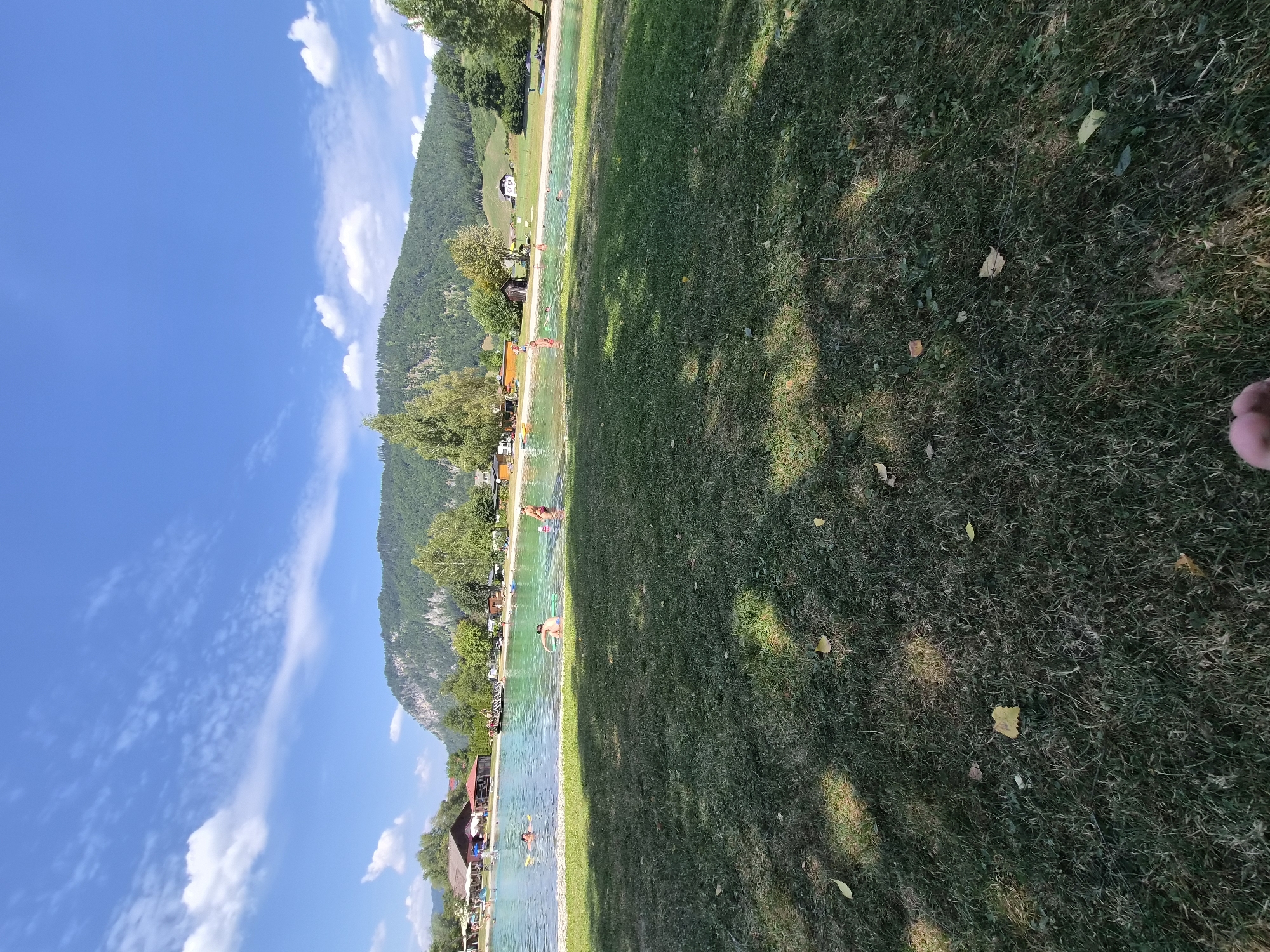 The image shows a panoramic view of a public swimming area on a sunny day. The foreground is dominated by a grassy lawn with scattered yellow leaves. Beyond the lawn, a clear, turquoise body of water is visible, with several people swimming and playing. In the background, there are buildings that appear to be part of the recreational facility, including structures that resemble restaurants or changing rooms. A lush, tree-covered mountain rises behind the swimming area, with a clear blue sky dotted with white clouds above. The scene suggests a popular summer destination. The location is identified as a bathing area in Landl, Austria.