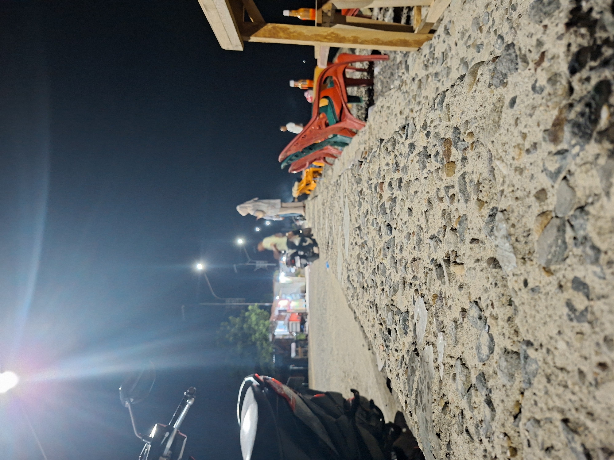 The image depicts a night scene in Lhokseumawe, Indonesia, where a row of street food vendors is set up along a sidewalk. The foreground is dominated by a rough concrete wall, with a motorcycle parked near it. Beyond the wall, there are food stalls illuminated by overhead streetlights, with indistinct figures of vendors and customers visible around them. The stalls display colorful plastic chairs stacked on top and bottled drinks. The background is a dark night sky, with streetlights casting a glow.