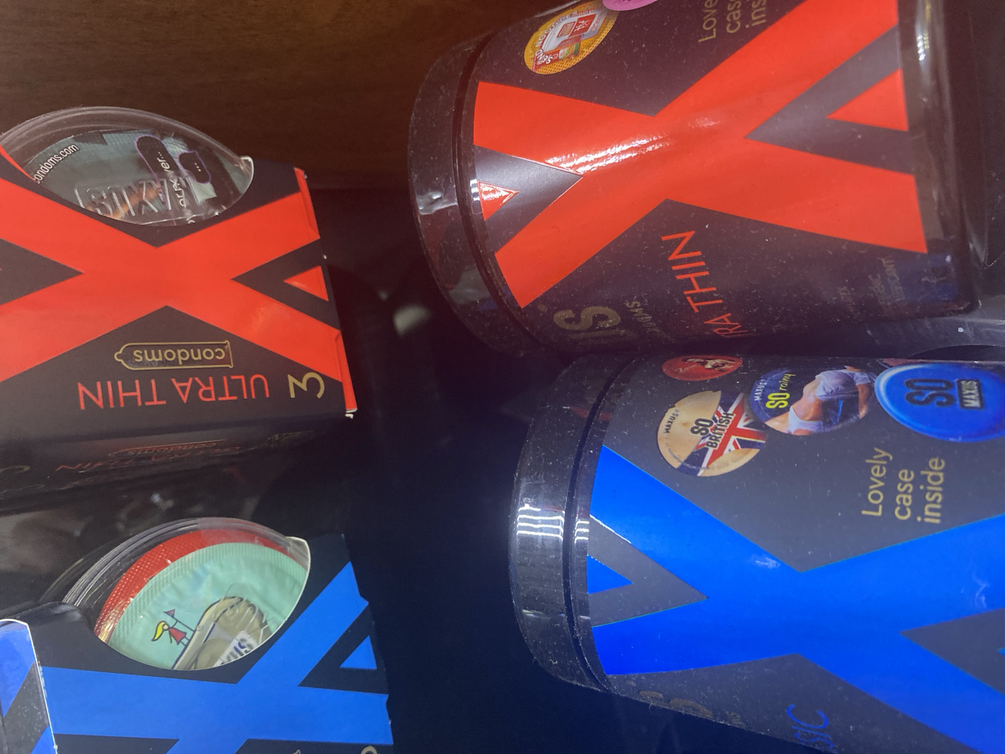 The image shows a close-up view of several packages of condoms. One package, rectangular in shape, has the words "ULTRA THIN" and "condoms" visible, with a large red "X" across the front. Another package is cylindrical and also features a large red "X" and the words "ULTRA THIN". A third package, also cylindrical, has a blue "X" and the words "Lovely case inside," "SO rainy," "SO BRITISH," and "MAXUS." Some individual condoms in wrappers are visible at the top of the red X box and at the bottom of the blue X box. The image is taken in Chaikovsky, Russia.