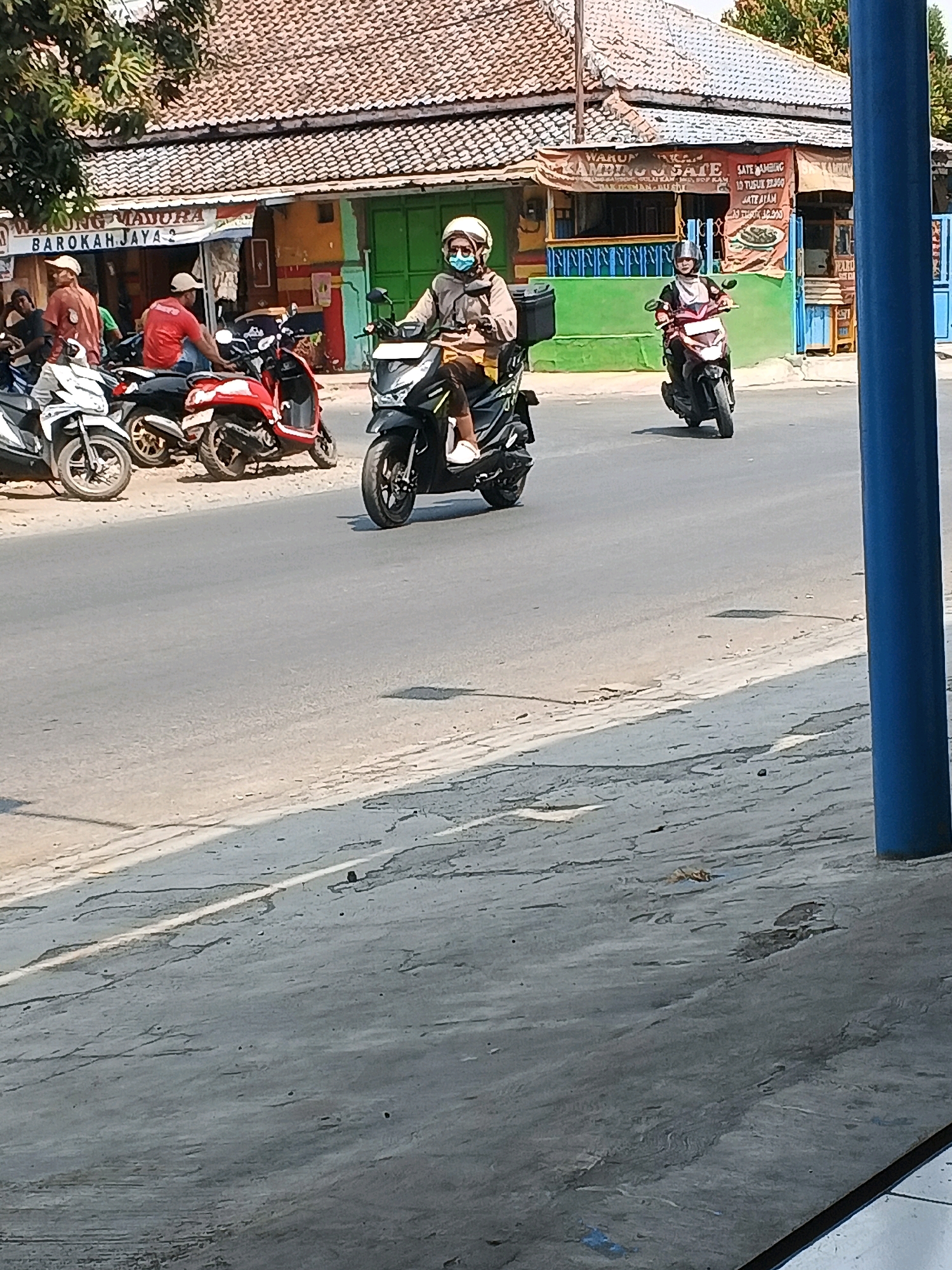 The image shows a street scene in Taman, Indonesia. A man wearing a helmet and a face mask is riding a black scooter, with a delivery box attached to the back. In the background, there are several buildings with signs, and other motorcycles are parked. The street is paved and appears to be a sunny day.