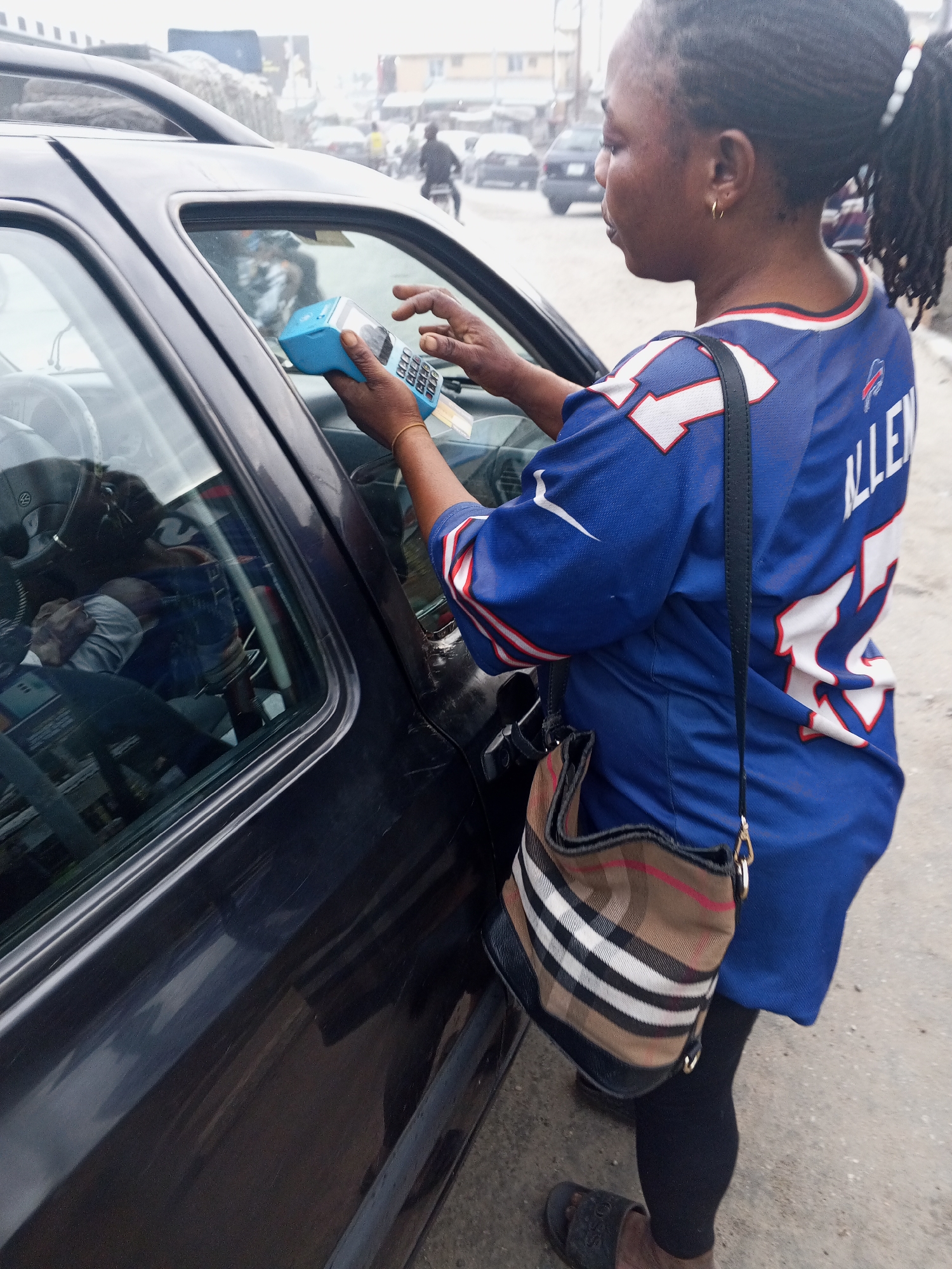 A woman, identified as a driver, stands next to a black car in Lokoja, Nigeria. She is wearing a blue football jersey and a plaid crossbody bag. In her hands, she holds a blue payment terminal, with her fingers positioned over the keypad, suggesting she is in the process of making or receiving a payment. The background shows a street scene with other vehicles and buildings, indicative of an urban environment. The overall scene appears to be a typical transaction occurring within the city.