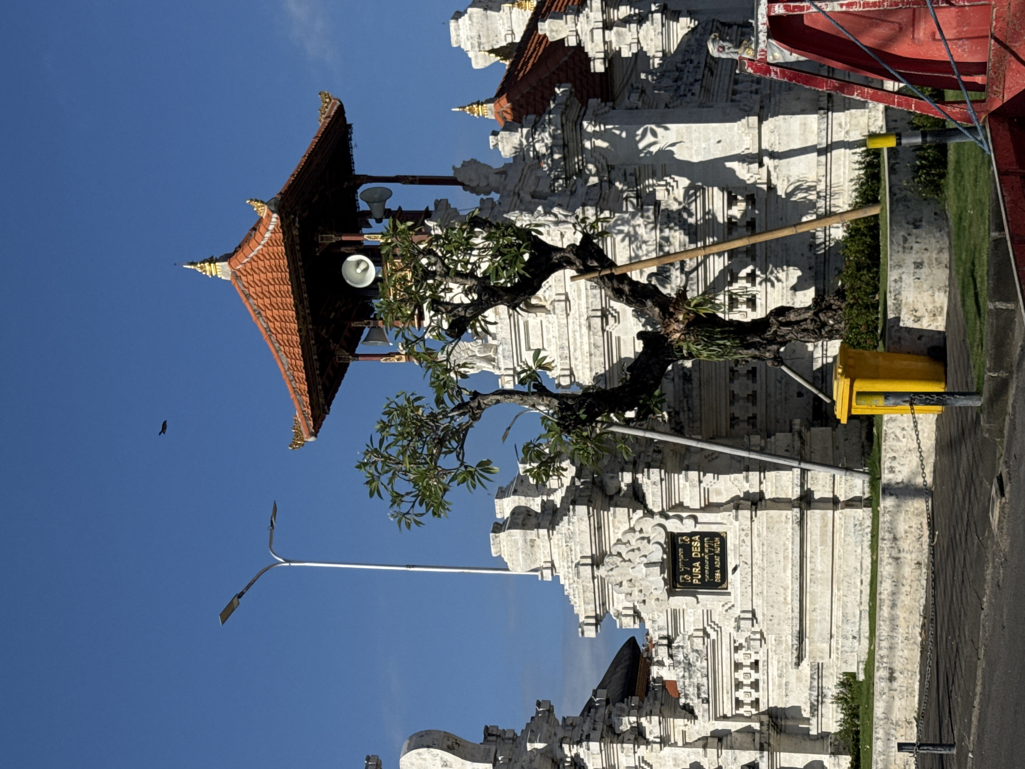 The image shows a Balinese temple in Kuta Selatan, Indonesia. The temple has intricate white stone carvings and a multi-tiered red roof. A tree grows in front of the temple. The sky is clear and blue. A bird is flying in the sky. A street lamp and a yellow object are also visible. A sign on the temple reads "Pura Desa".