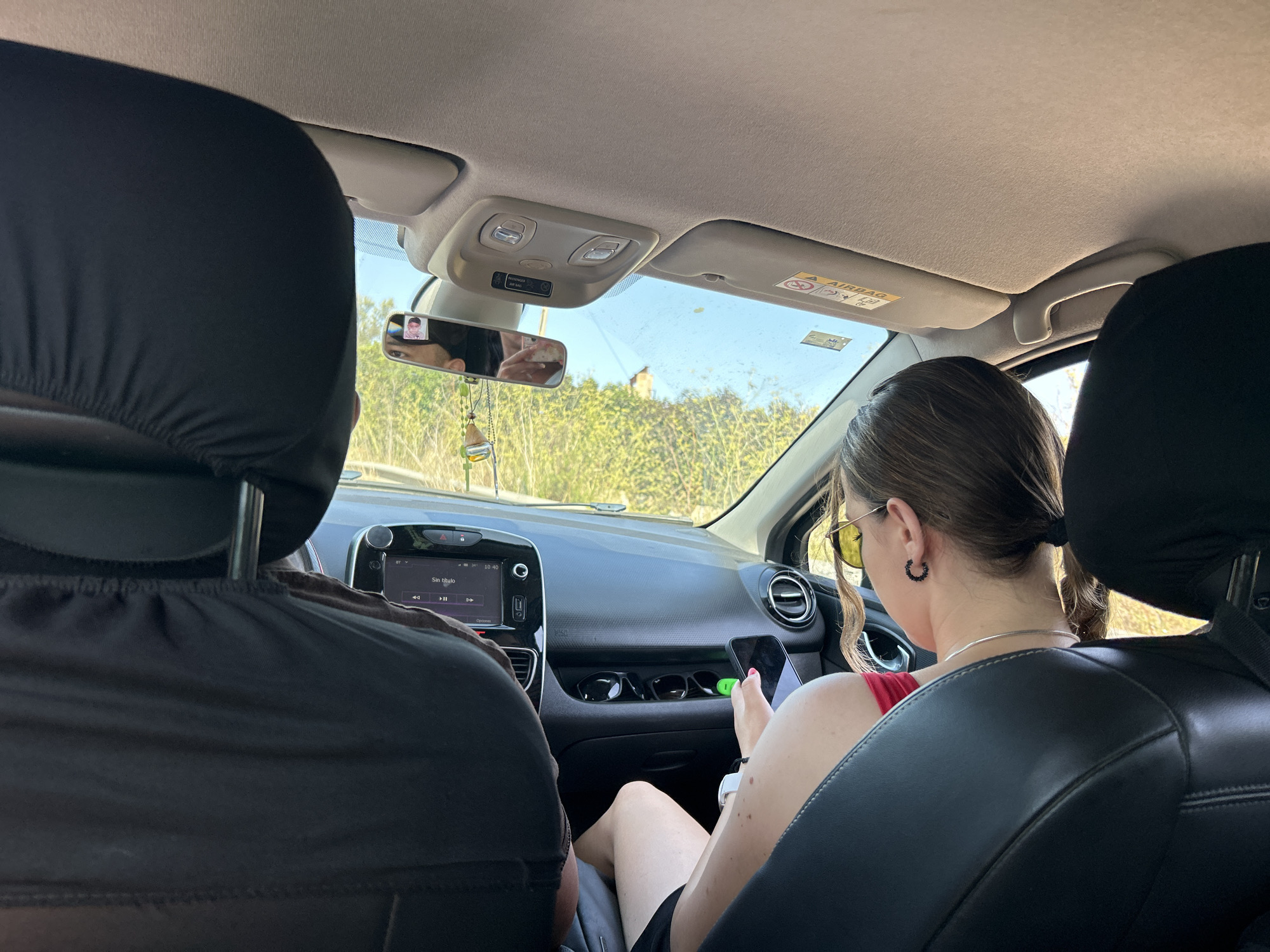 The image shows the interior of a car, viewed from the back seats. A person in the passenger seat, a young woman with brown hair tied in a ponytail, is looking down at her phone, which she holds in her right hand. She is wearing a red top, black shorts, sunglasses, and large hoop earrings. The driver, whose back is to the camera, is wearing a black shirt. The car's dashboard is visible, with a touch screen display showing "Sin titulo". The car is moving, as indicated by the greenery outside the windows, suggesting a road trip. Based on the style of the car and the dashboard display, it appears to be a modern hatchback. The ambient lighting and the greenery outside are consistent with a Mediterranean climate, possibly during daytime. Although the exact location is not specified in the image, if this scene were to be placed in Capdepera, Spain, the surrounding vegetation would likely consist of pine trees, olive groves, and Mediterranean scrubland, characteristic of the island of Mallorca. The road would likely be paved, with possible dry stone walls or agricultural terraces visible in the distance. The sky would be a clear blue, common in this region during the summer months.