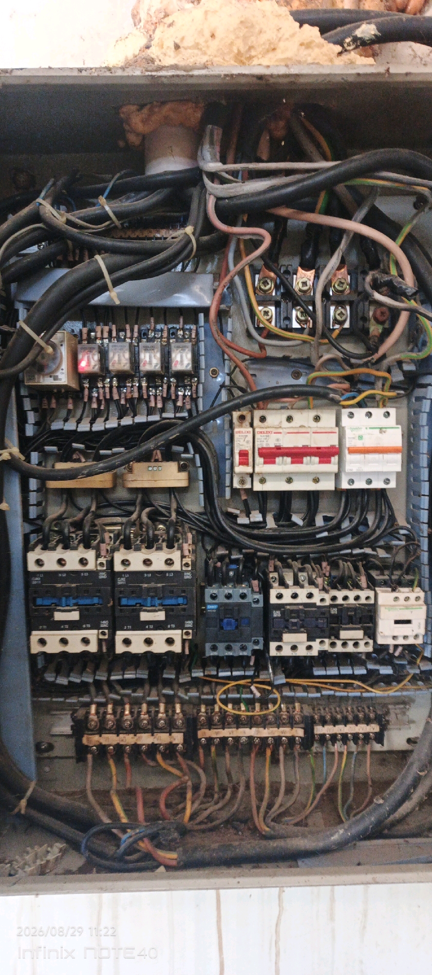 The image shows an electrical control panel with numerous wires, relays, and circuit breakers. The panel appears to be dusty and somewhat disorganized. There are several rows of relays, a few larger contactors, and a strip of circuit breakers. Some wires are neatly bundled with ties, while others are loose. The overall impression is of an industrial or commercial electrical system. The location is identified as Patampanua, Indonesia.