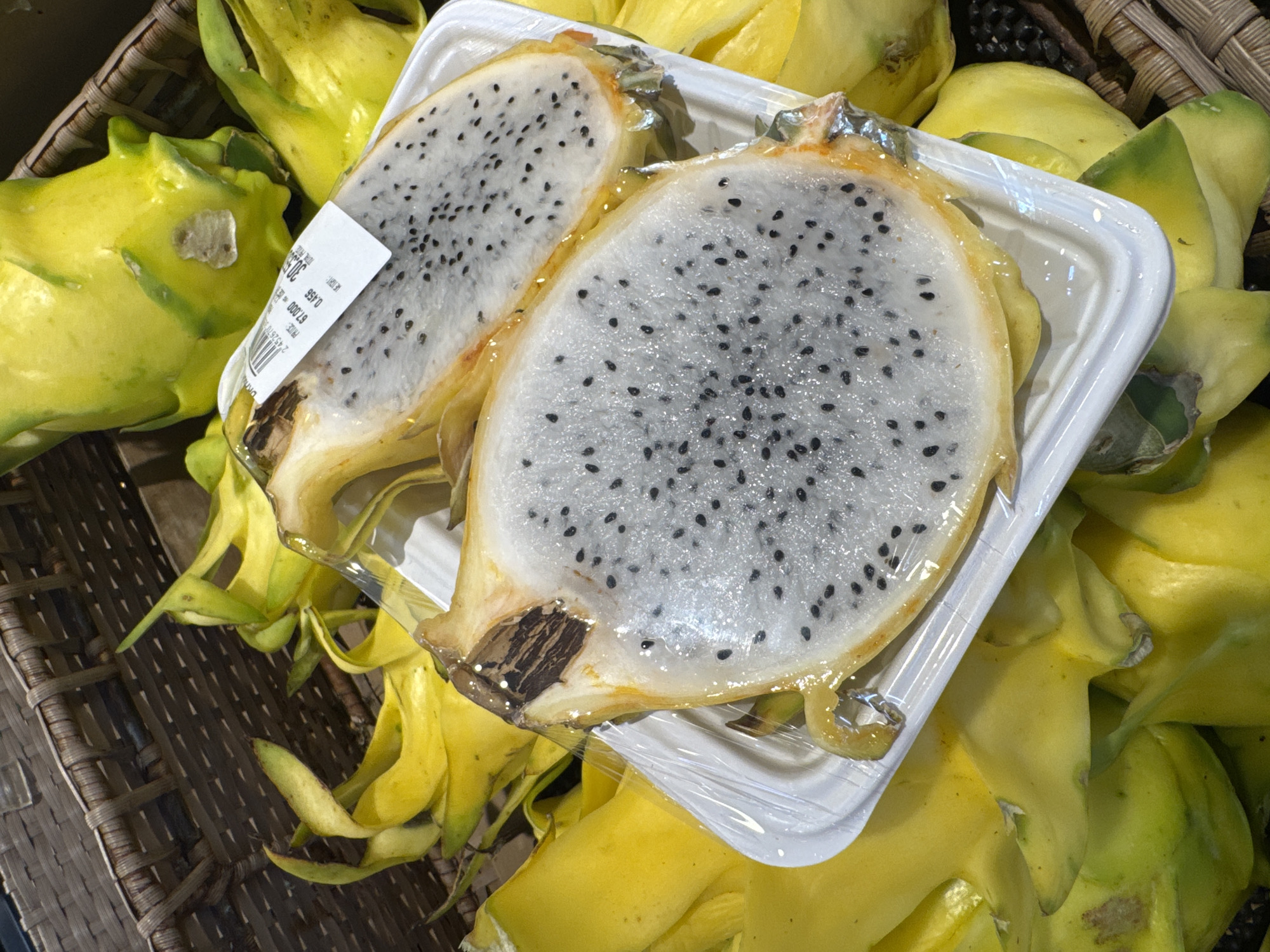 The image shows two slices of yellow dragon fruit, each cut in half, packaged in clear plastic wrap on a white polystyrene tray. The tray is placed on top of a pile of whole, yellow dragon fruits inside a brown wicker basket. The location is inferred to be Kuta Selatan, Indonesia, based on the subject matter (yellow dragon fruit) being a fruit commonly found in tropical regions like Indonesia.