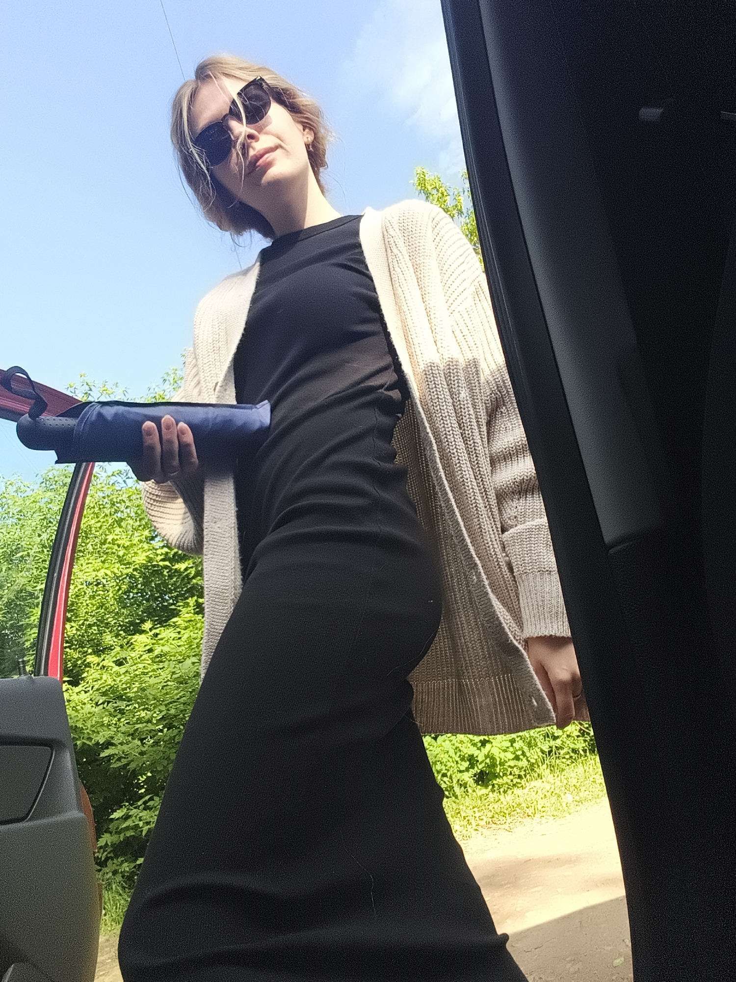The image shows a woman standing next to a car, likely outside of it. She is wearing a black dress, a light beige cardigan, and sunglasses. She is holding a dark blue object, possibly a compact umbrella. The background shows green foliage and a blue sky. A red car door is visible to the left of the woman, and part of the interior of the car is on the right of the frame. The location is Izhevsk, Russia.