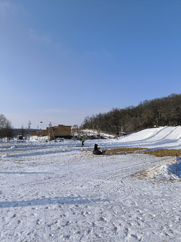 The image displays a snow-covered landscape in Prybuzke, Ukraine, under a clear blue sky. In the mid-ground, a snow-covered field shows numerous tracks from foot traffic and sleds. To the left, several brick-colored buildings are visible behind a low white fence, alongside utility poles and power lines. Further right, a densely forested hill rises, with a prepared snow tubing or sledding slope featuring multiple parallel tracks descending towards the foreground. A parking area with vehicles is situated between the buildings and the slope. Two individuals are present: one person, wearing a bright green or yellow jacket, stands upright in the mid-ground, facing away from the viewer. Another person is seated on a dark-colored inflatable snow tube, facing left, with a pink item visible near them. A blue mesh safety net is partially visible on the far right edge of the foreground. The overall scene suggests a winter recreational area.