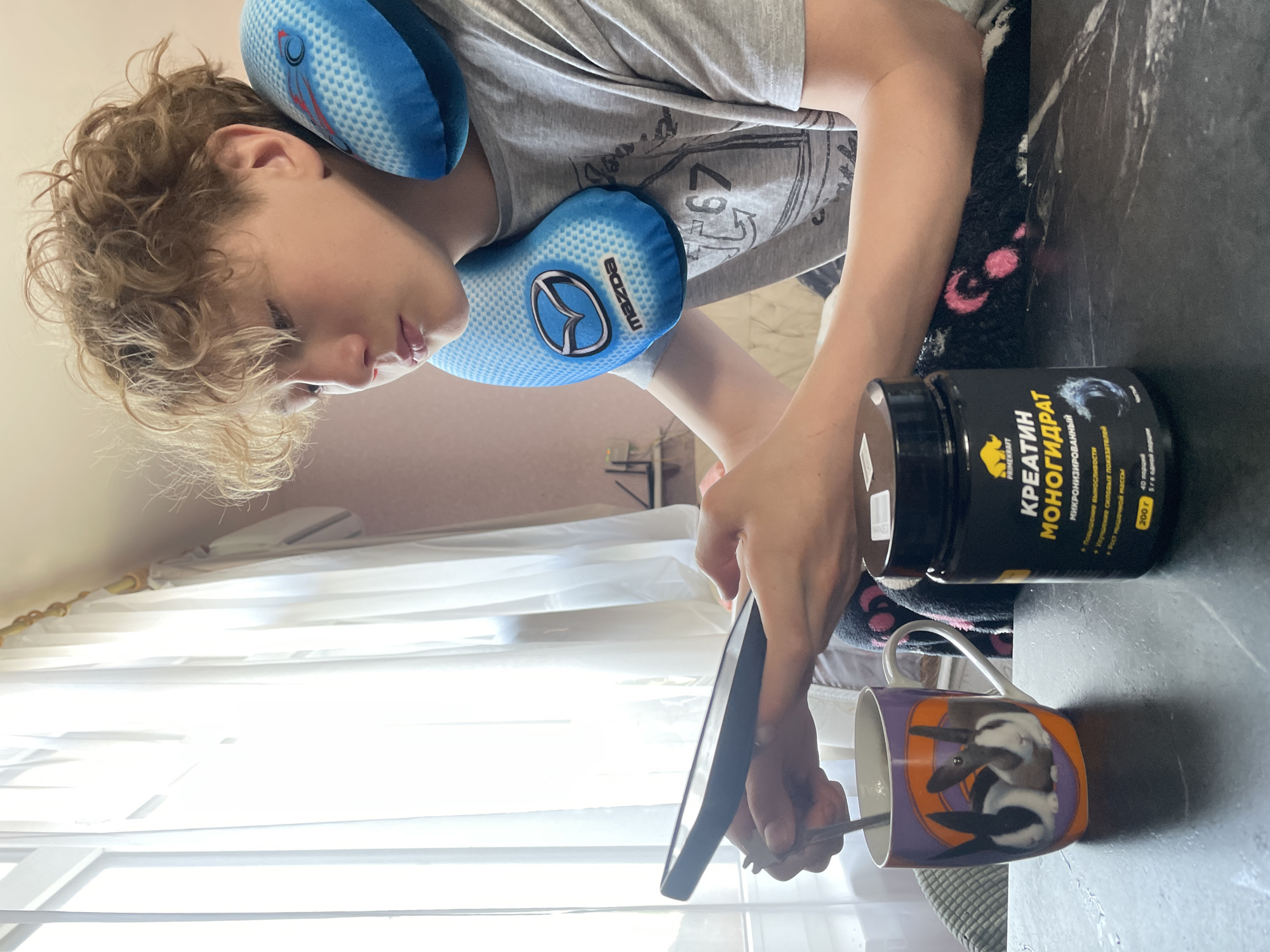 A young person with curly blonde hair, wearing a grey t-shirt and a blue neck pillow with the Mazda logo, is standing next to a table. On the table is a black container labeled "КРЕАТИН МОНОГИДРАТ" (Creatine Monohydrate), a mug with bunny designs, and the person is holding a spoon and what appears to be a phone. The photo is taken indoors, with a window and light curtains visible in the background. There is no verifiable information to confirm the location as Baranovka, Russia.