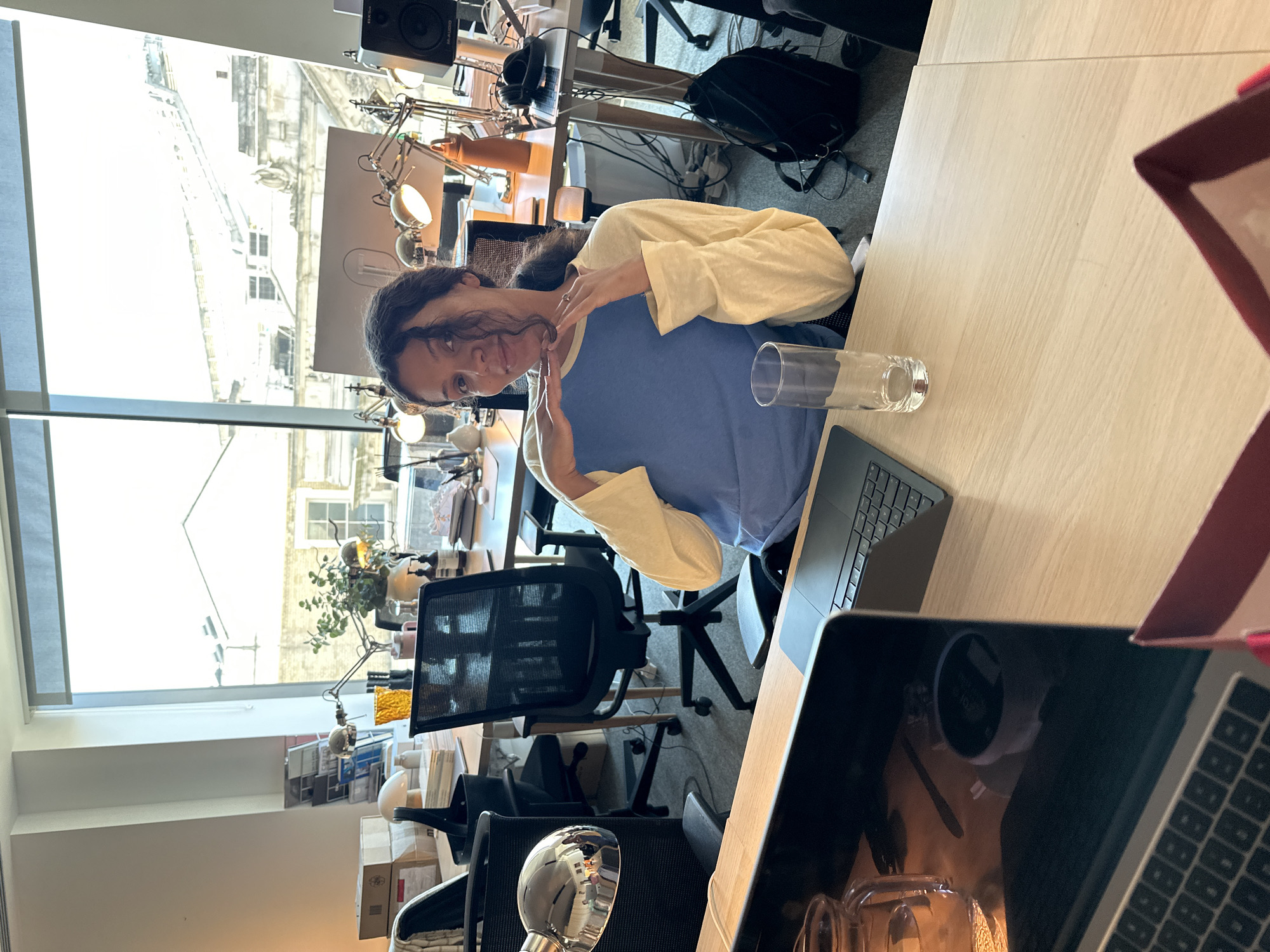 The image shows a woman seated at a desk in a brightly lit office space. She is wearing a blue and cream-colored top and has dark hair. She appears to be interacting with the camera, making a gesture with her hands near her face. There is a glass of water on the desk near a laptop. The office has multiple desks, chairs, and lamps visible. A large window provides a view of an urban landscape. The buildings visible through the window appear to be consistent with the architecture of London, United Kingdom. The window allows daylight to stream into the space.