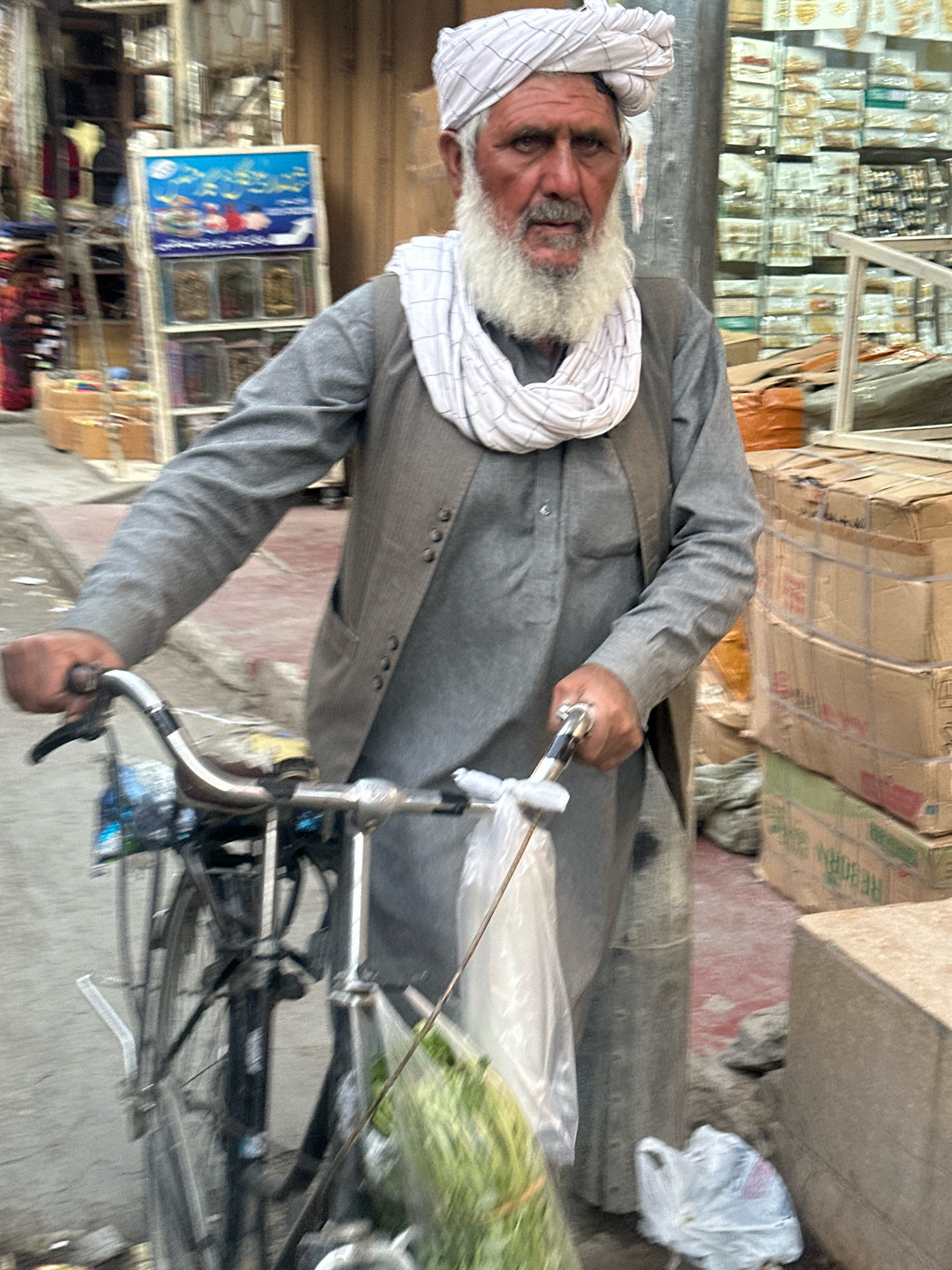 An older man with a white turban and white beard is standing in a street, holding the handlebars of a bicycle. He is wearing a grey tunic and a darker grey vest. His left hand is resting on the handlebar, and his right hand is holding a small bag. The bicycle has a front basket containing a plastic bag with what appears to be green produce. In the background, there are shops with shelves displaying various items, including boxes stacked high and some signage. The ground is a mixture of dirt and red pavement. The overall scene suggests a bustling marketplace environment in Quetta City Tehsil, Pakistan.