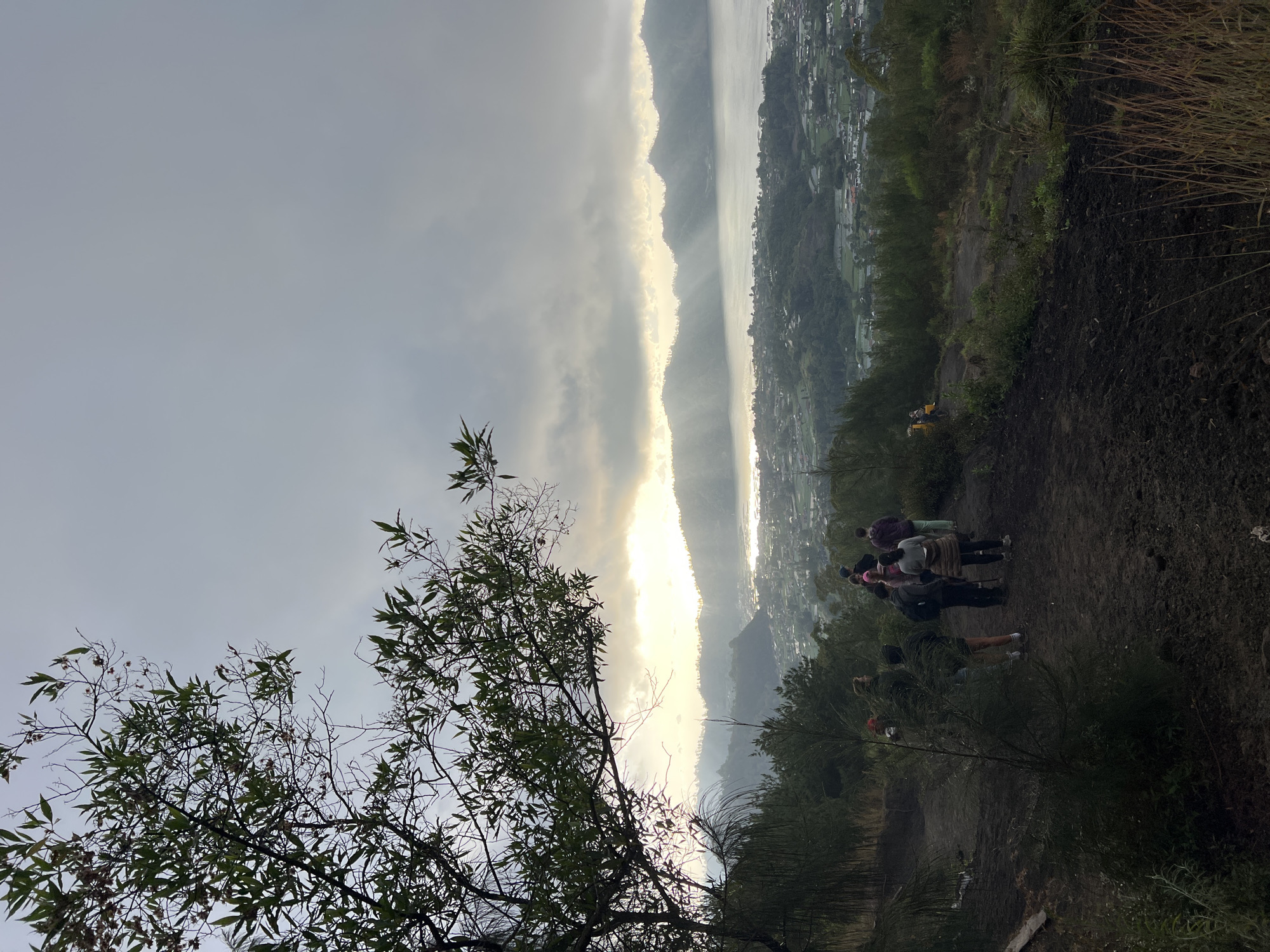The image depicts a group of people on a hiking trail overlooking a lake and a town, likely in or around Kintamani, Indonesia. The time appears to be early morning, given the low-angle sunlight breaking through the clouds and illuminating the mist over the water. The foreground shows a dirt path winding uphill, with sparse vegetation and some larger trees on the left. A group of approximately eight individuals, dressed in casual hiking attire, are visible on the path, some walking and others paused. They are facing away from the camera, looking out at the panoramic view. In the mid-ground, a vast body of water, presumably a lake, is visible, bordered by hills and a town with numerous buildings spread across the landscape. The background consists of layered hills or mountains, with the sunbeams creating a dramatic effect as they pierce through the overcast sky. The overall atmosphere is serene and suggestive of a dawn hike in a scenic natural environment.