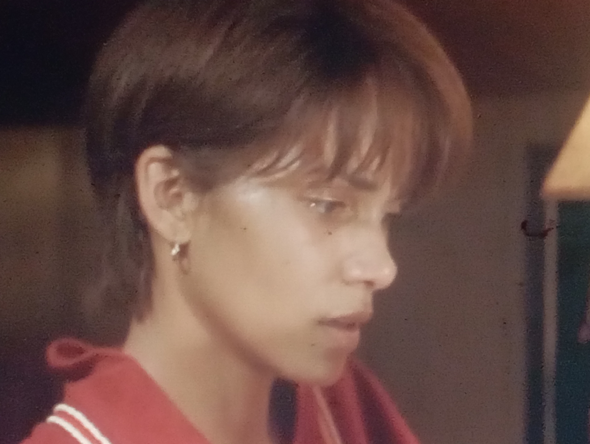 The image is a close-up, medium-shot photograph of a young person, possibly female, with short, dark brown hair styled in a mullet. The person is wearing a red top with white stripes on the collar and a small hoop earring in their left ear. Their head is turned to the right, and their gaze is directed downwards and to the right, with their lips slightly parted. The lighting is dim and warm, suggesting an indoor setting, possibly at night. The background is dark and out of focus, with a hint of a lamp or light fixture on the right side. There is no discernible connection to the city of Sofiivska Borshchahivka, Ukraine, as no landmarks or contextual elements indicate this location. The photograph appears to be a candid shot or a still from a video, exhibiting grain and a slightly soft focus.
