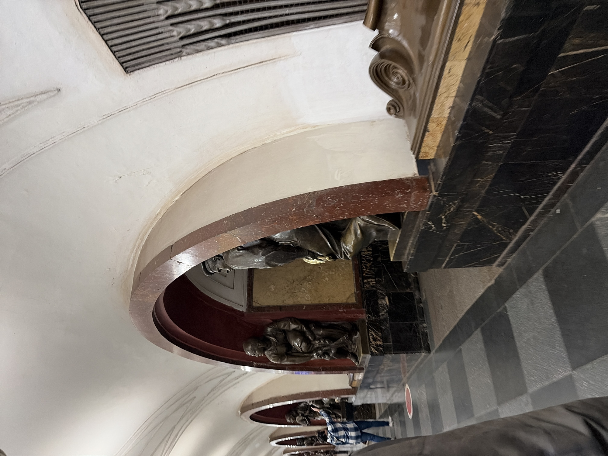 The image shows a section of a Moscow Metro station. In the foreground, a person in a dark jacket is partially visible on the left. To the right, the station features a series of arched alcoves, each containing a dark bronze statue. The statues depict figures in historical attire, some holding tools or objects. The statues are mounted on black marble pedestals. The station's architecture includes curved white ceilings and reddish-brown arched frames around the alcoves. The floor is paved with dark and light gray square tiles. In the background, more statues and arches are visible, receding into the distance. A person in a blue and white checkered shirt is seen standing in one of the alcoves, gesturing. The lighting appears to be artificial, typical of an underground subway station.