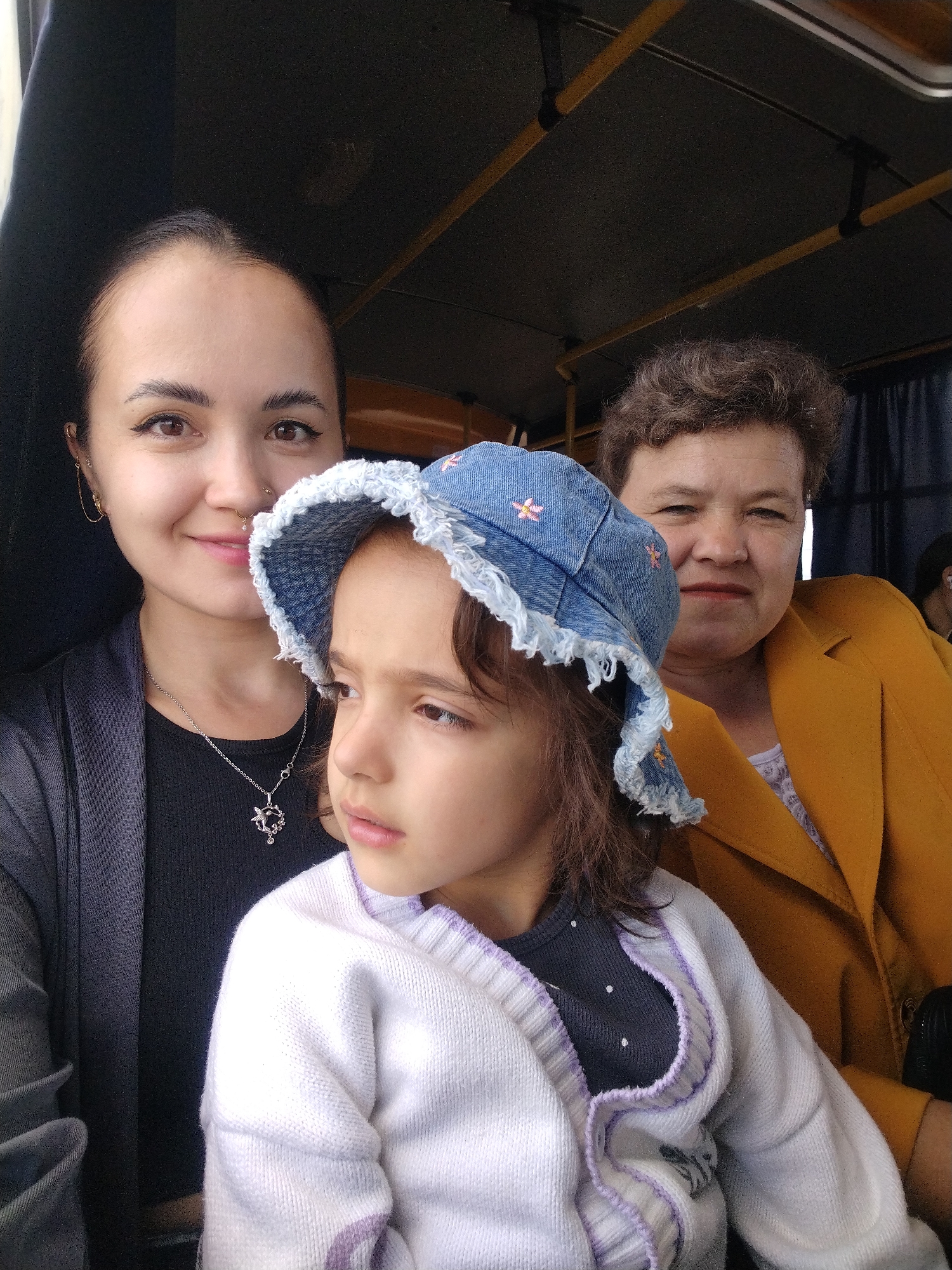 The image shows three people inside a vehicle, possibly a bus or a van. In the foreground, a young girl wearing a denim hat with frayed edges and a white cardigan is seated. To her left, a woman with dark hair pulled back and wearing a dark jacket over a black shirt is visible. She is smiling and wearing a necklace. To the right of the girl and behind her, an older woman with short, curly hair is wearing a mustard-yellow jacket over a white top. The interior of the vehicle appears utilitarian with yellow handles and a dark ceiling. The lighting suggests it is daytime. The location is identified as Noviy, Russia.