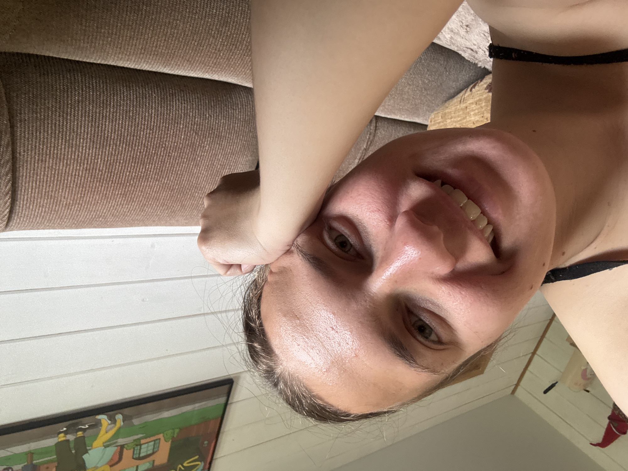 A selfie of a woman with light skin and short, graying hair is angled with her head positioned towards the right side of the frame. Her face is well-lit, revealing her smile and her brown eyes. Her hand is positioned near her forehead. A black choker is around her neck.
Behind her is a couch and wooden wall, and a framed picture hanging on the wall. Based on the context, the location is potentially Раздолье, Russia.