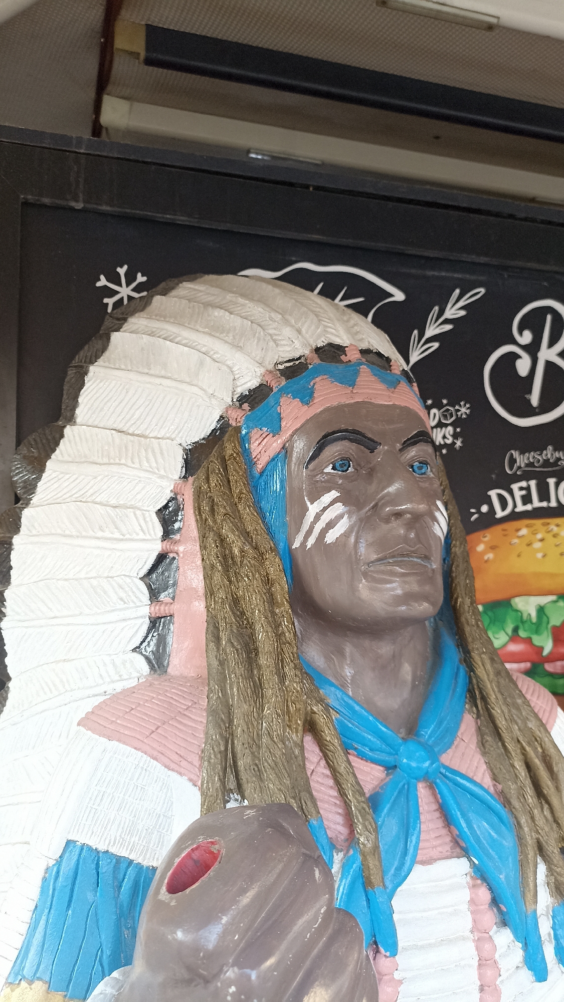 A vibrant, multi-colored statue of a figure resembling a Native American chief is prominently displayed in this eye-level, close-up shot. The figure has dark brown skin, piercing blue eyes, and white ceremonial paint lines on its cheeks. It wears an elaborate feathered headdress, primarily white with black accents, featuring pink and blue bands around the forehead. Long, textured brown hair or dreadlocks frame the face, and a bright blue necktie or scarf is worn around the neck. The figure’s attire includes a pink and white patterned garment. In its left hand, part of an object with a red interior, possibly a ceremonial pipe, is visible. The statue conveys a stoic and contemplative expression.
The figure is positioned against a dark background that appears to be a chalkboard or menu board. White chalk-like drawings and text are visible on this surface. Notable text includes a large, ornate "B" at the top right, and partially visible words like "Cheeseburg" (likely "Cheeseburger"), "DELIC" (likely "DELICIOUS"), and "DOO KS" (possibly "DRINKS"). A snowflake-like asterisk symbol and stylized leaf patterns are also drawn on the board. Below the text, a blurry image of a hamburger is discernible. Above the board, a light-colored, textured ceiling and a black metal beam are visible, indicating an indoor or sheltered commercial setting.