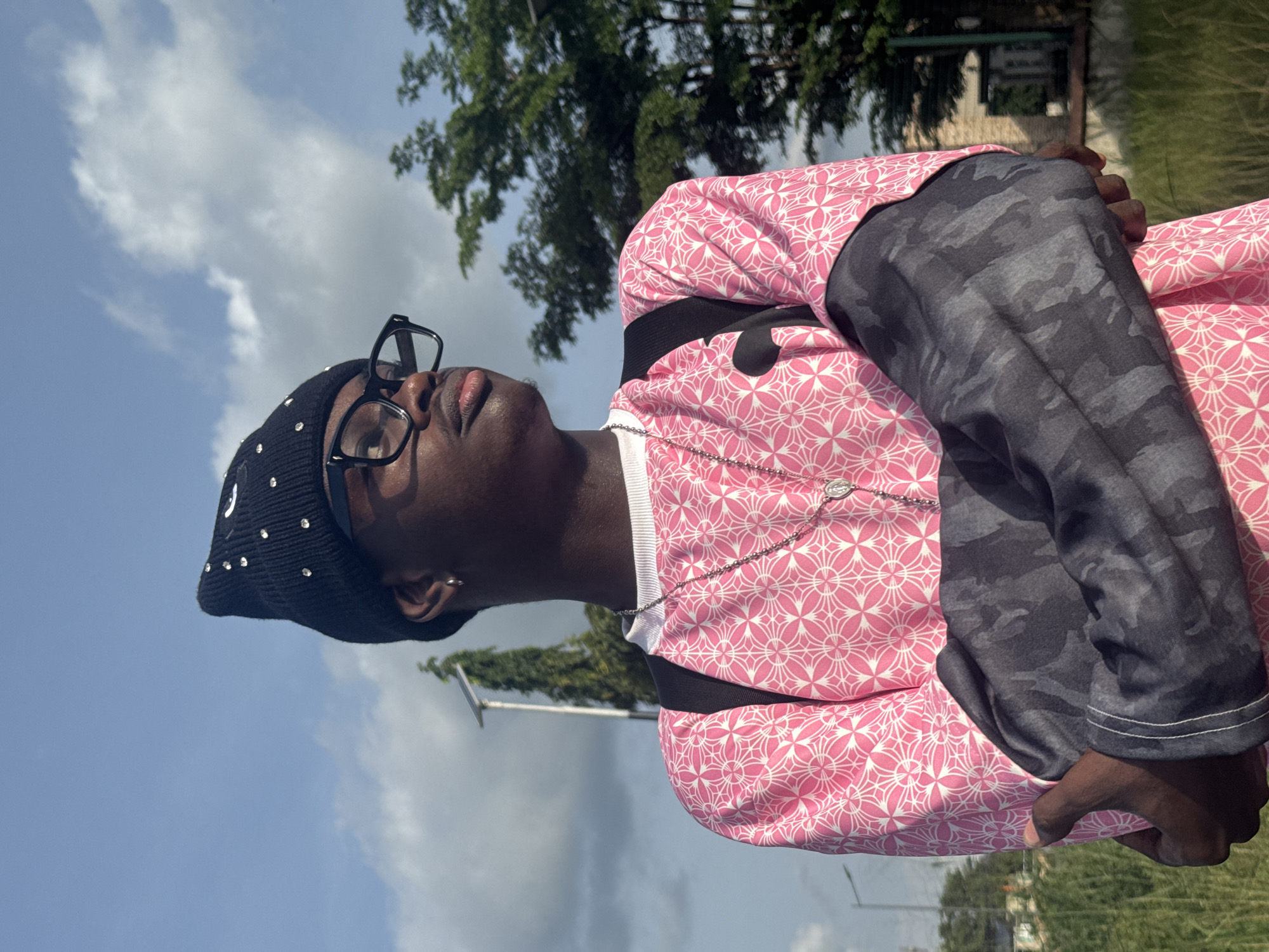 A person with dark skin is outdoors in Ihiagwa, Nigeria, looking up at the sky. They are wearing glasses, a black knit cap with silver decorations, a silver chain necklace, a pink patterned shirt, and a gray camouflage-patterned long-sleeved shirt underneath. The person has their arms crossed. Behind them are trees, a blue and cloudy sky, a light post, and a building in the distance.