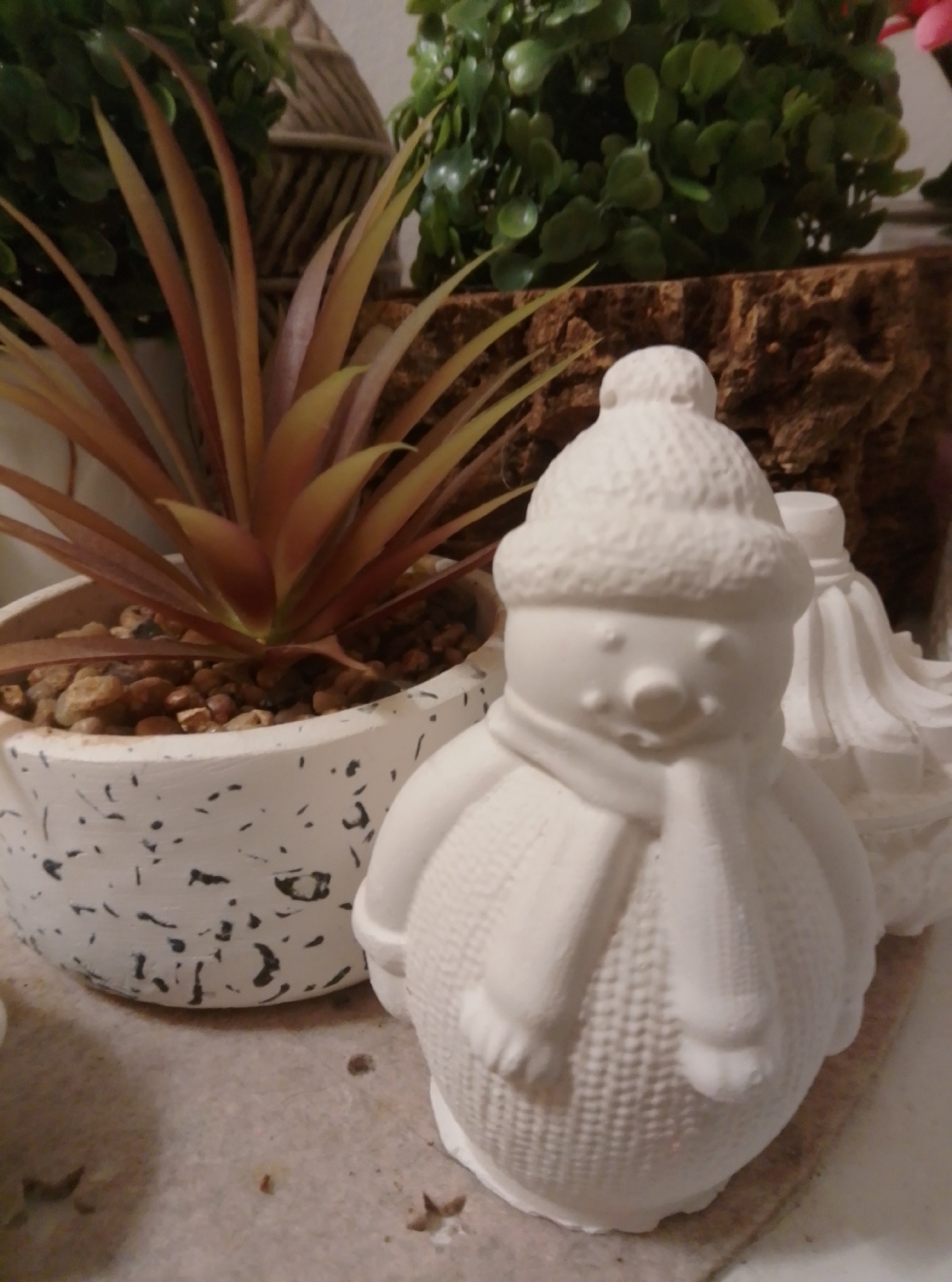 The image shows a close-up of a ceramic snowman figurine in the foreground, wearing a knitted hat and scarf. Behind the snowman, there is a potted plant with spiky, reddish-brown leaves in a speckled white pot filled with small stones. In the background, there are decorative green plants and a textured brown material, possibly bark or wood. The overall scene appears to be a still life or decorative arrangement. The image does not contain any identifiable landmarks or elements that would suggest a specific location in the city of Wedel, Germany.