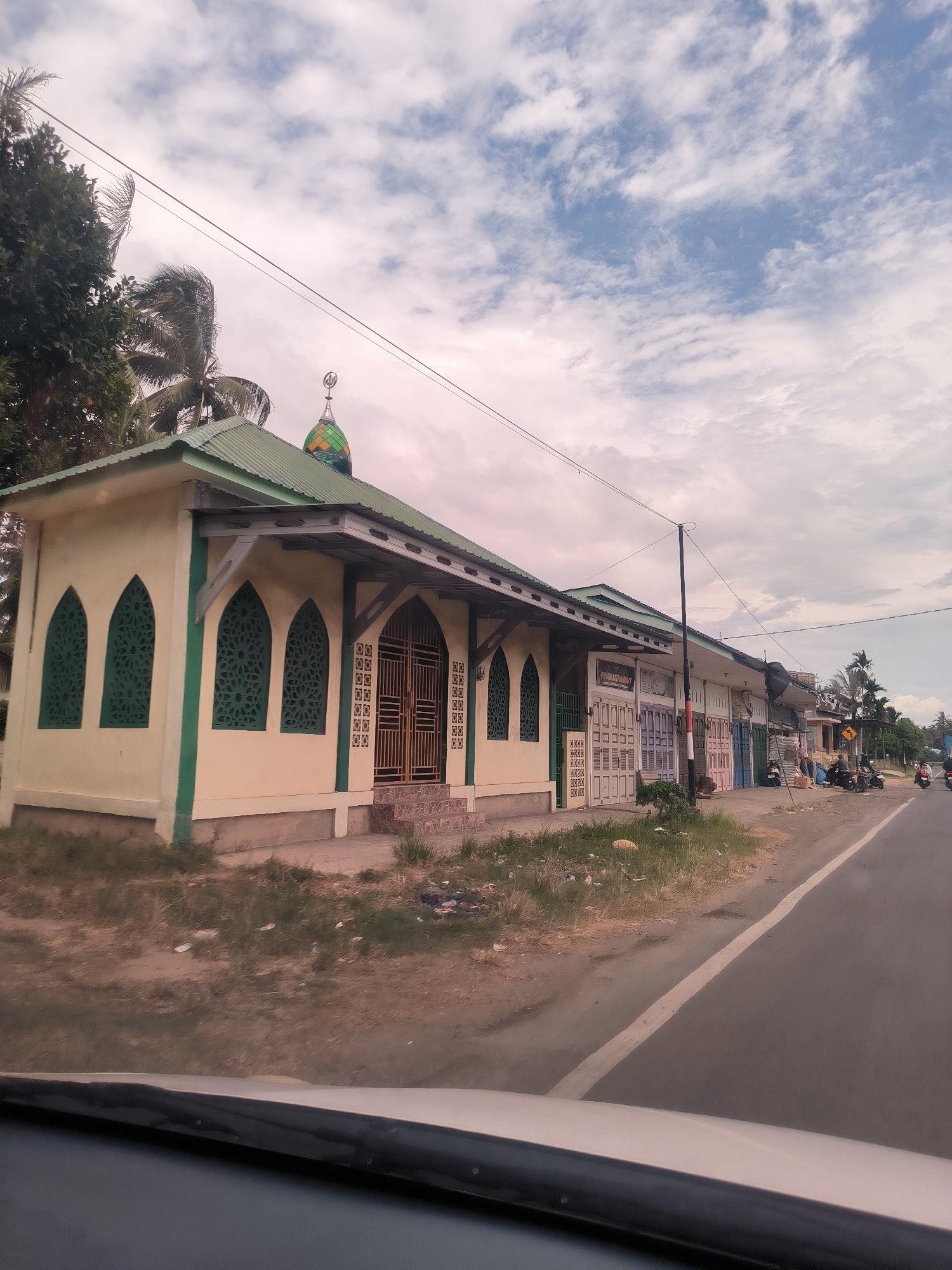 This image presents a roadside view captured from within a vehicle, with part of its dashboard visible at the bottom. The scene depicts a street in what appears to be a semi-urban or rural area of Pandrahkandeh, Indonesia, under an overcast sky filled with scattered white and grey clouds.
On the left side of the frame, a distinctive, single-story building resembling a mosque or prayer house dominates. It features light cream-colored walls with a dark green trim, a sloped green corrugated metal roof, and a small, colorful dome at the back, adorned with green, yellow, and orange patterns and topped with an Arabic symbol. The building's front has several arched window openings with dark green, ornate lattice screens, and a prominent double door with similar decorative metalwork. Behind this building, lush green trees, including palm trees, are visible, with power lines stretching across the sky.
To the right, a row of simple commercial buildings or shops extends along the road. These structures have various colored metal roll-down shutters, some closed, and a common design. One shop clearly displays the Indonesian text "PANGKAS RAMBUT," indicating it is a barbershop. Motorcycles are parked intermittently along the side of the asphalt road in front of these shops. A utility pole stands near the shops, and a yellow triangular warning sign is visible further down the road.
The road itself is dark grey with a single white dividing line. The lighting suggests it is daytime, possibly late morning or afternoon, given the brightness despite the cloud cover. The overall impression is of a quiet, functional streetscape in a community.