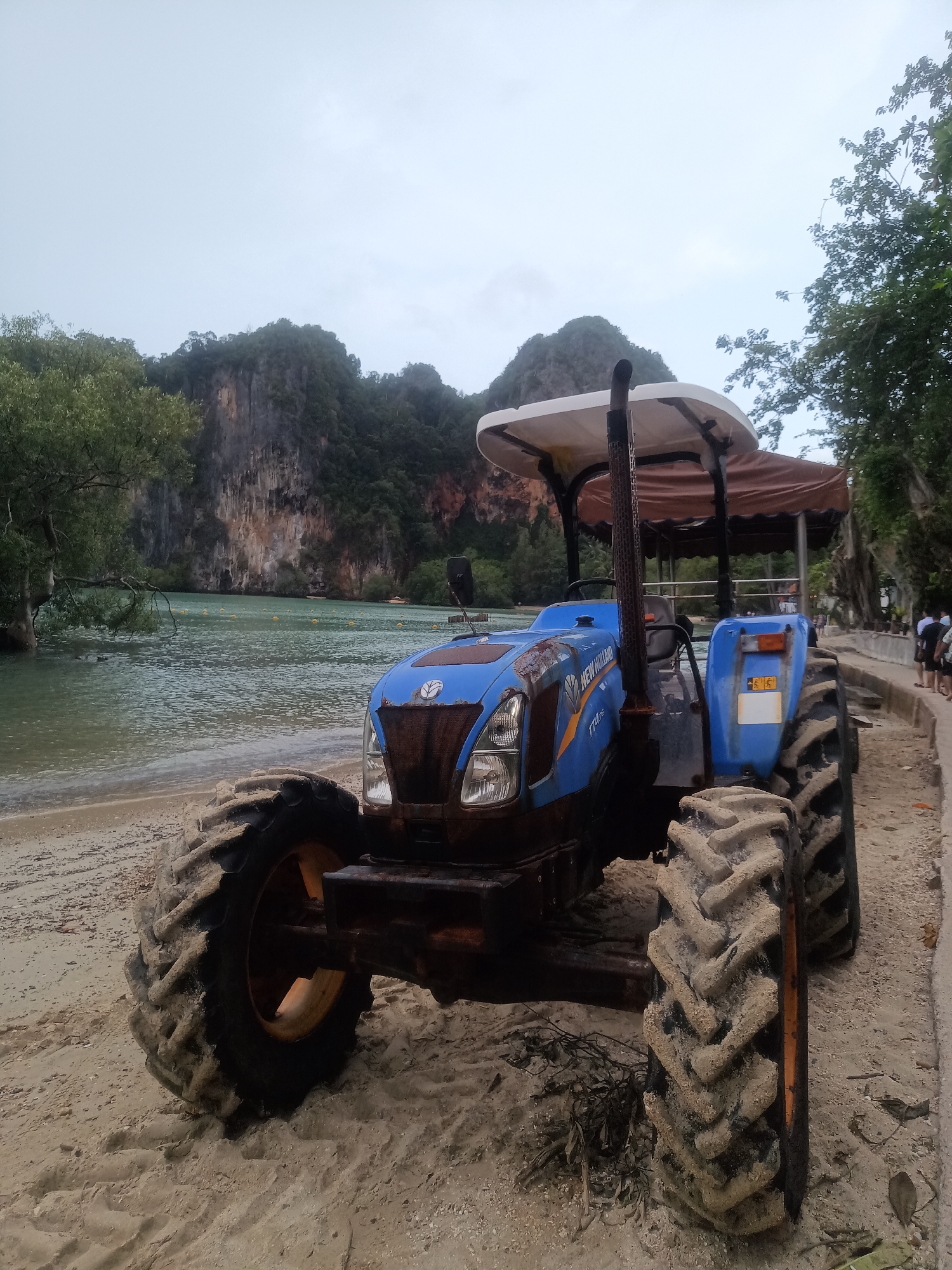 The scene depicts a blue New Holland TT4.75 tractor parked on a sandy beach. The large, knobby tires are covered in sand. The tractor has a white sunshade with a brown fabric lining. The tractor is located near a body of water, possibly a lagoon or bay, with limestone cliffs and tropical vegetation in the background. Some people can be observed on the right-hand side of the image. The image was captured on the beach, presumably in the city of Ban Ao Nang, Thailand. The sky appears overcast and cloudy.